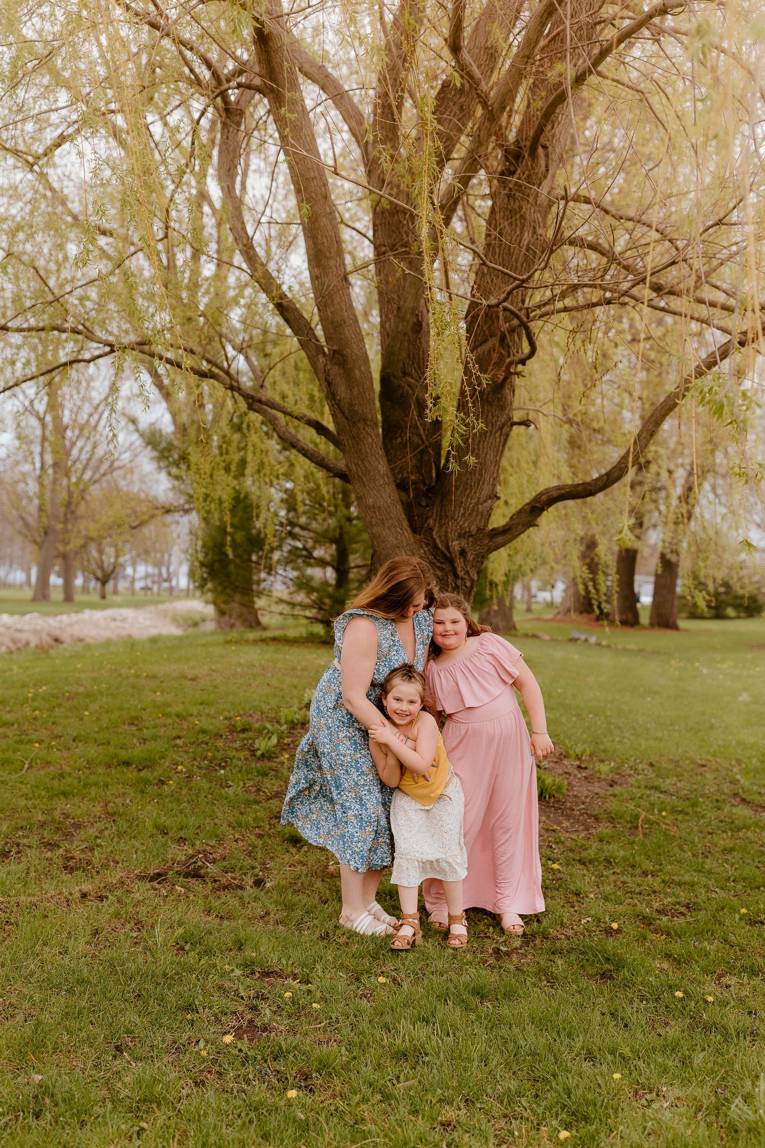 Three figures share a tender moment under a large willow tree in a park setting during golden hour.
