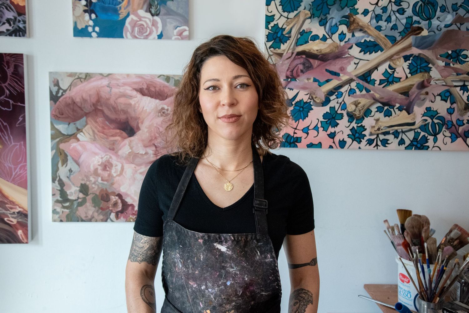 Artist in a black shirt works in their studio surrounded by colorful floral paintings and art supplies.