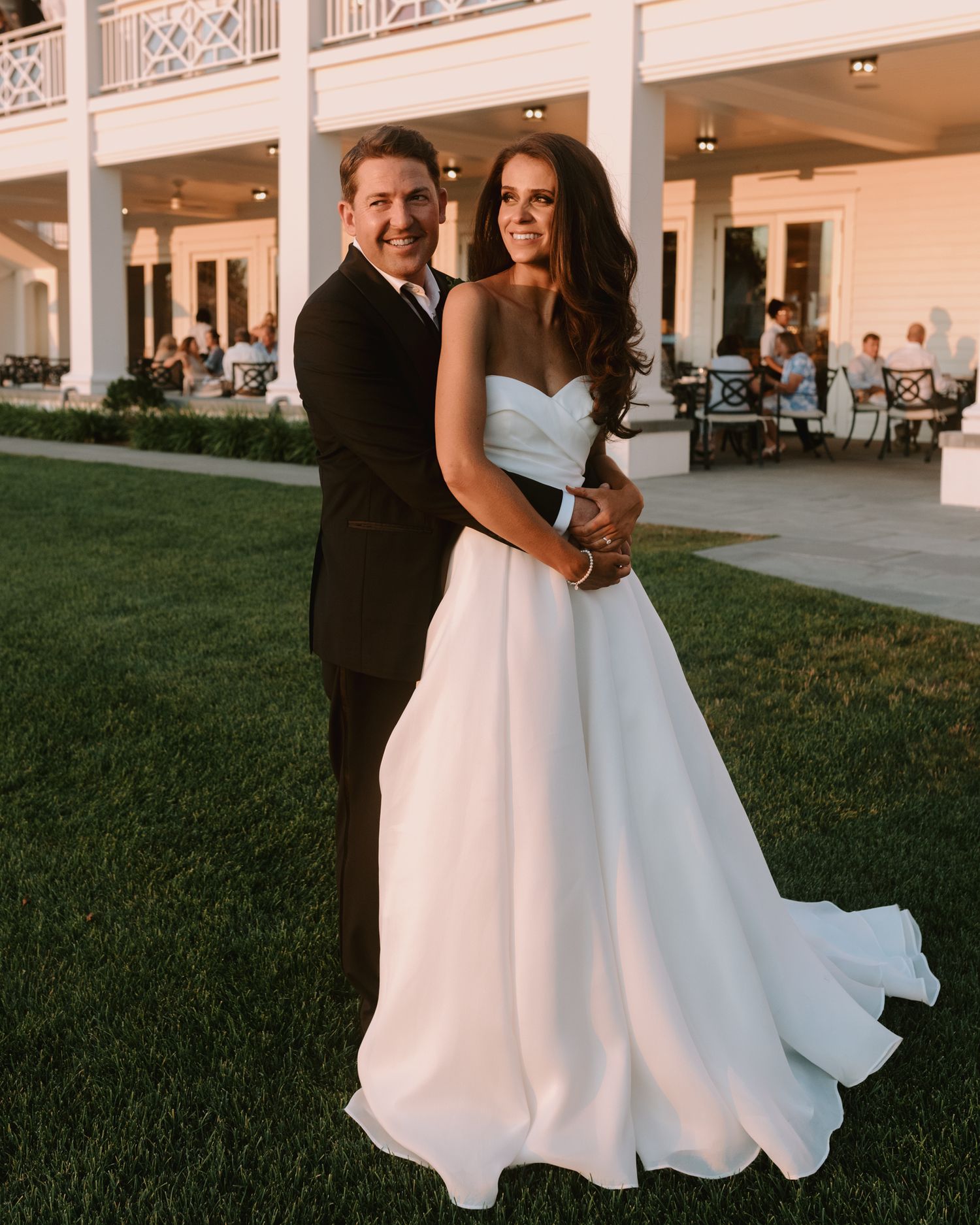A wedding couple poses together on the lawn in front of an elegant white colonial-style venue.