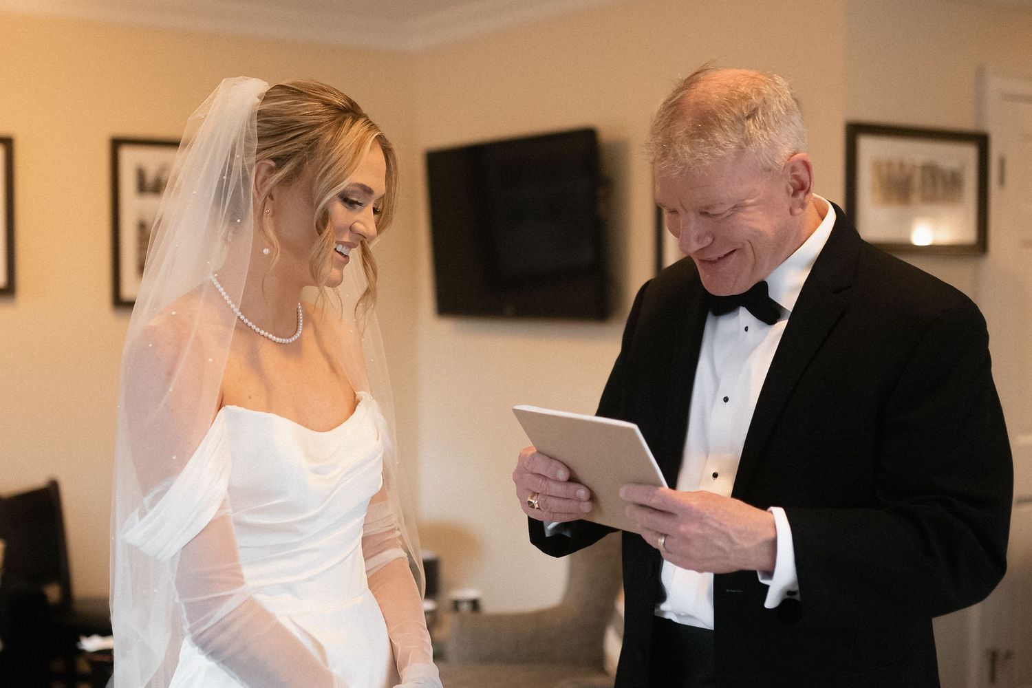 A bride in a white wedding dress and veil stands next to someone reading from a tablet in formal attire.