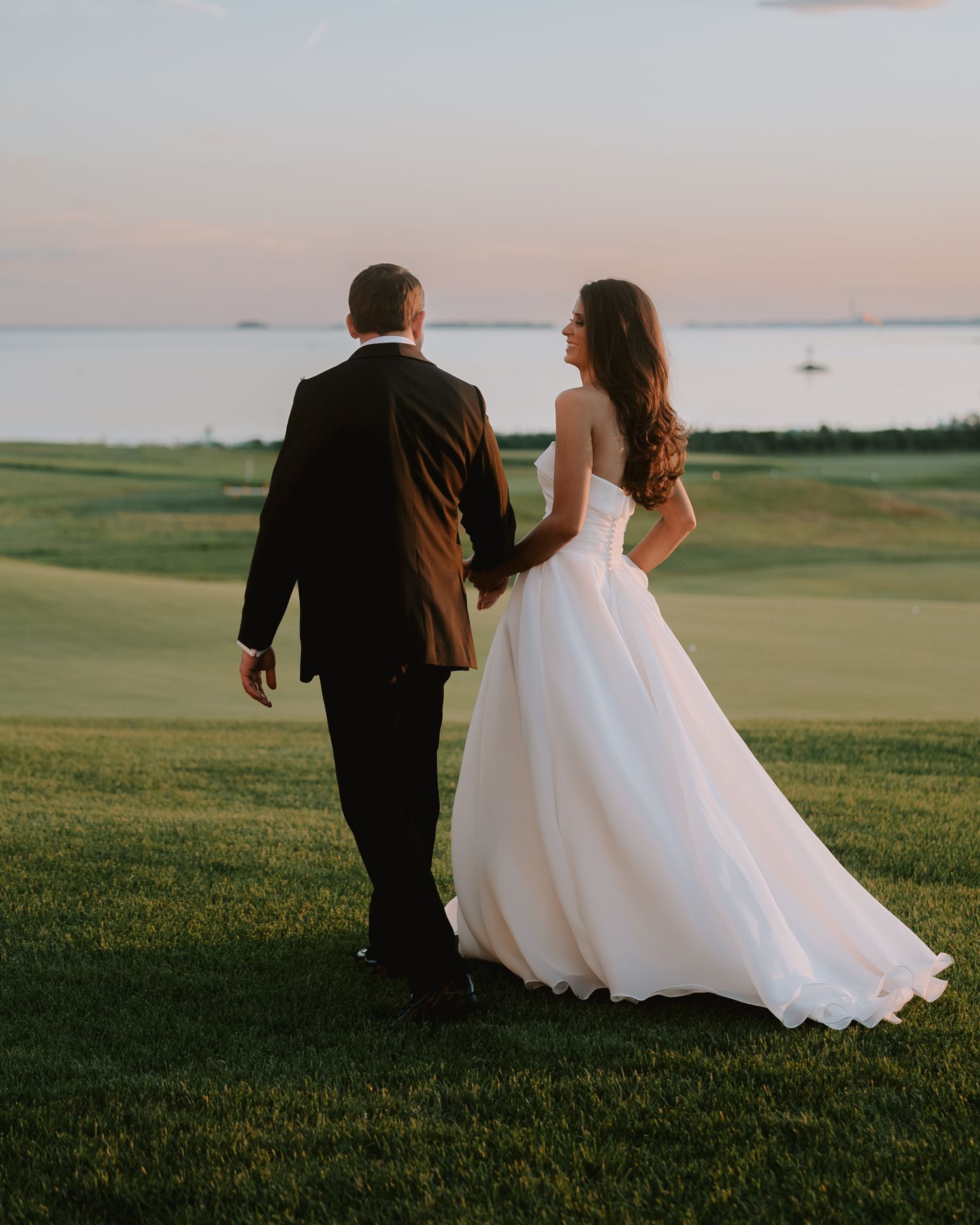 Couple walking across a lush green field in wedding attire during sunset.