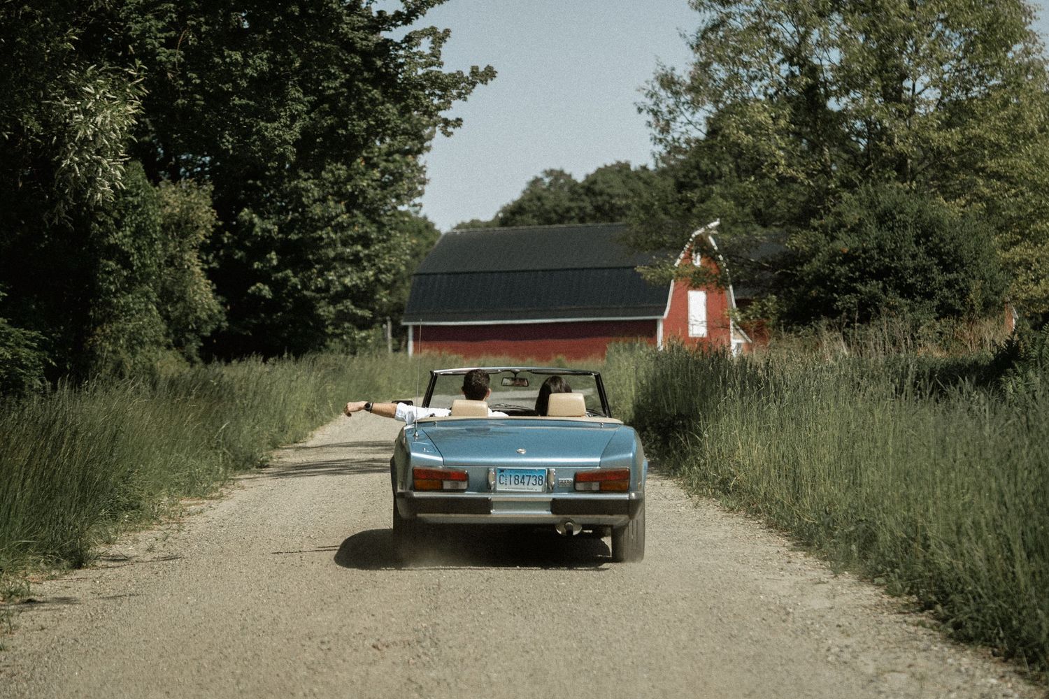 Vintage blue convertible car driving down a dirt path towards a red barn in summer.