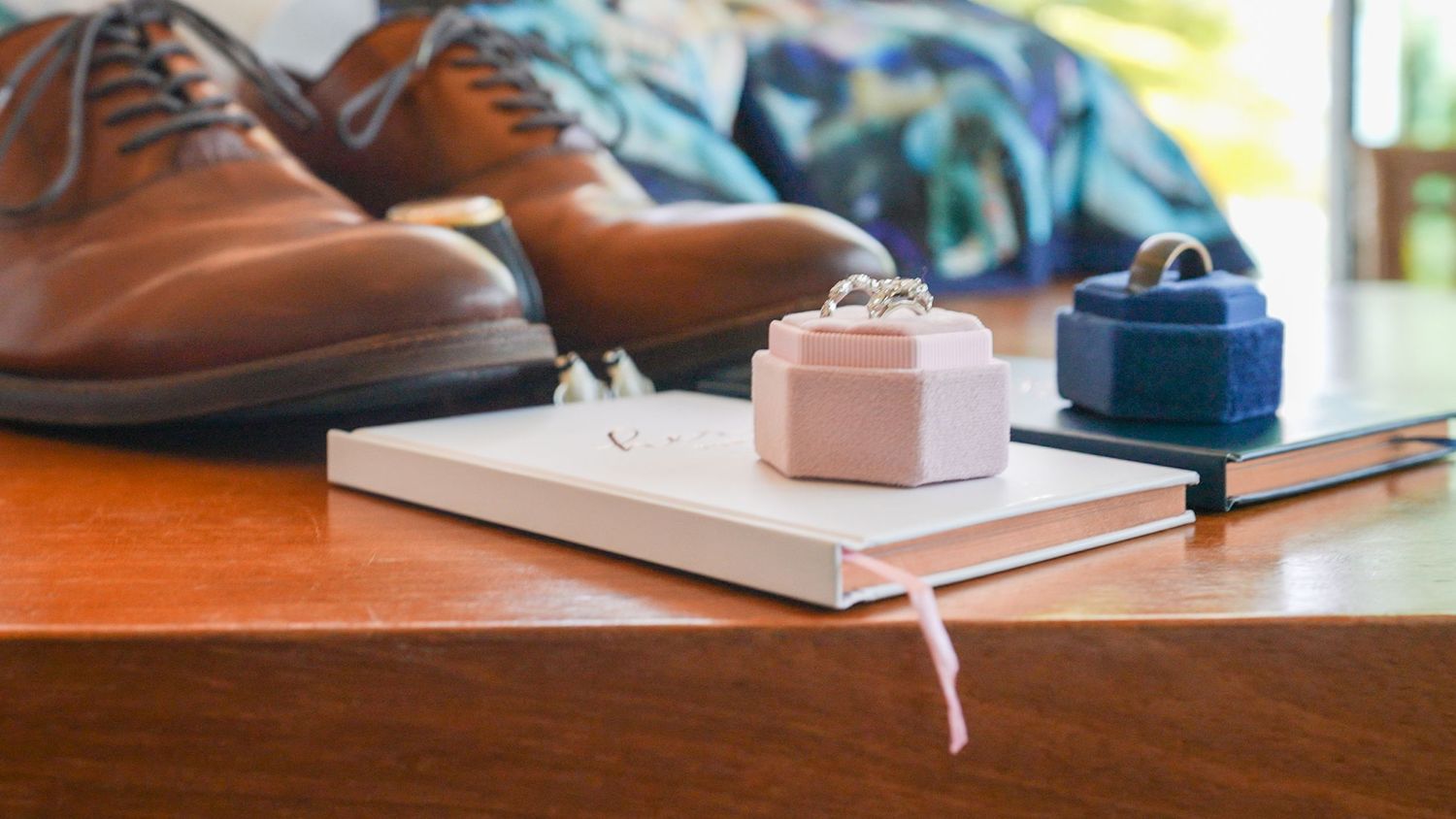 Brown leather dress shoes with wedding rings and gift boxes on wooden surface.