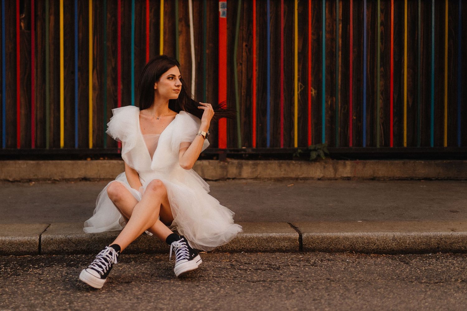 Person in a white dress and sneakers sitting against a colorful striped wall.