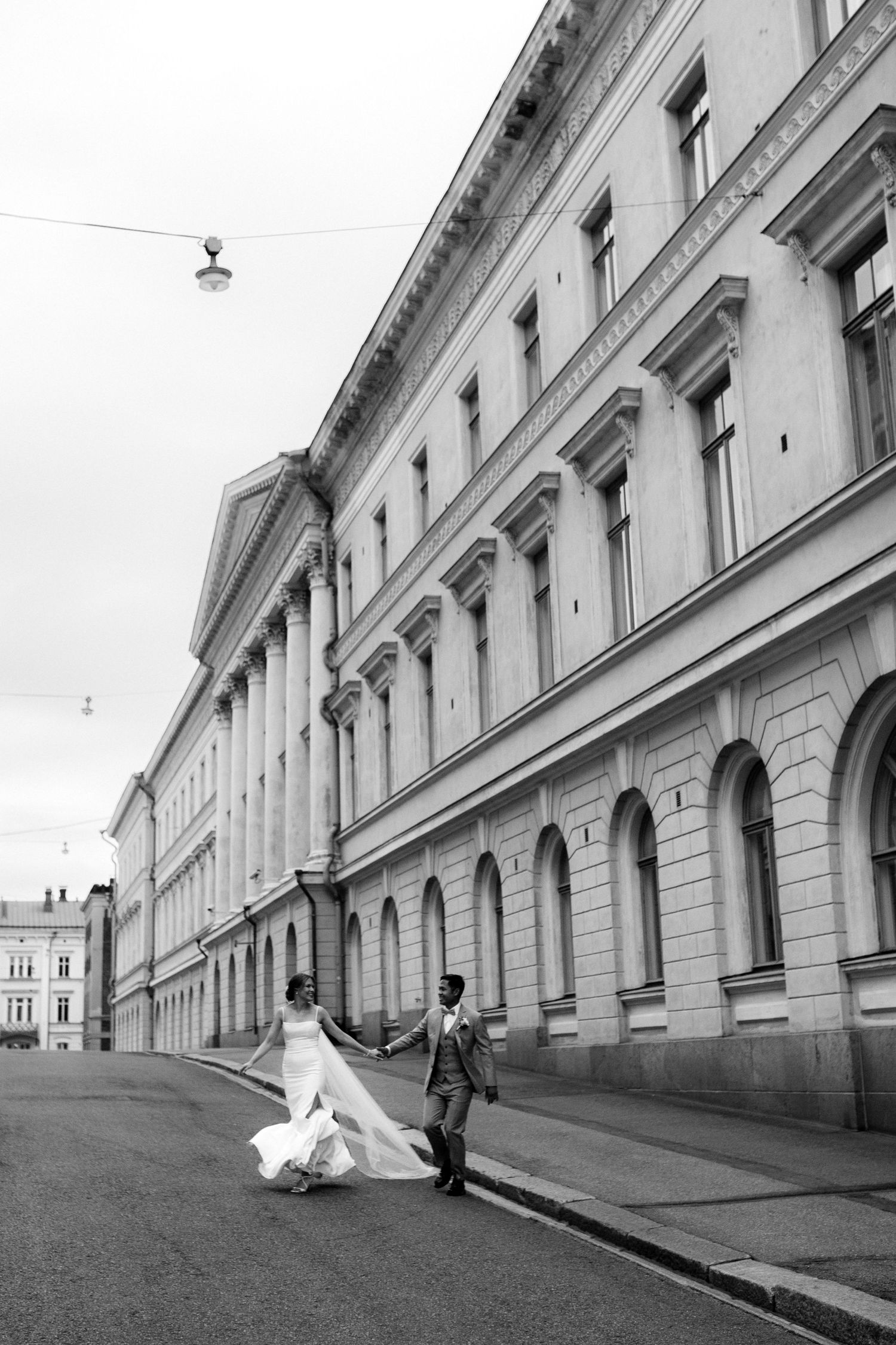 Mustavalkoinen, urbaani hääpotretti jossa hääpari juoksee kadulla kauniin rakennuksen edessä Kruununhaassa, Helsingissä.