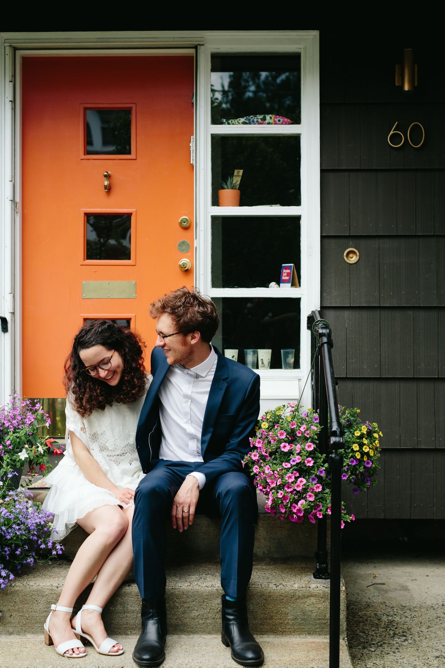 Bride and Groom hosting a backyard wedding reception share an intimate moment on steps in front of a bright orange door with purple flowers.