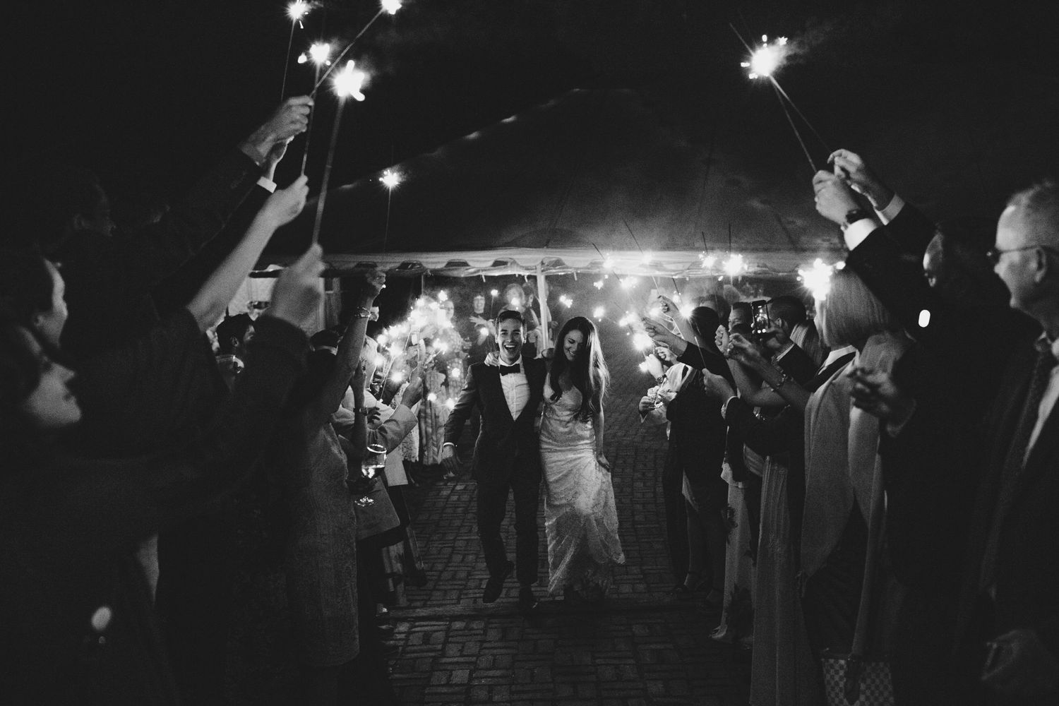 A black and white photograph shows a bride and groom walking under a arch of sparklers.