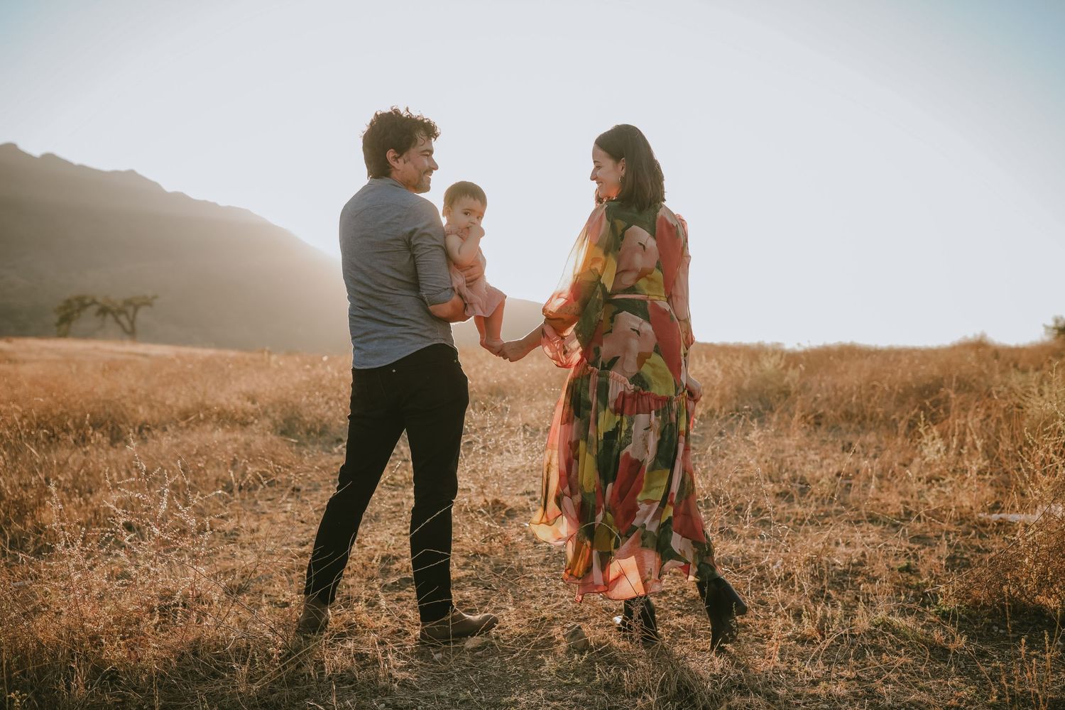 Three people dancing together in a golden field during sunset with mountains in the background.