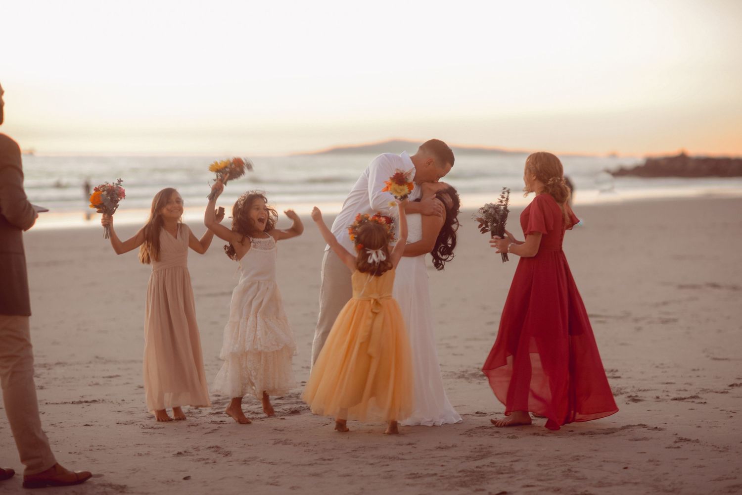 Group celebrating on beach at sunset wearing colorful formal dresses.
