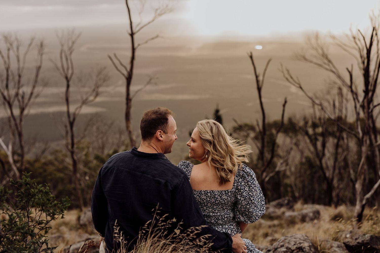 A couple shares an intimate moment while sitting among dry grasses with a scenic view.
