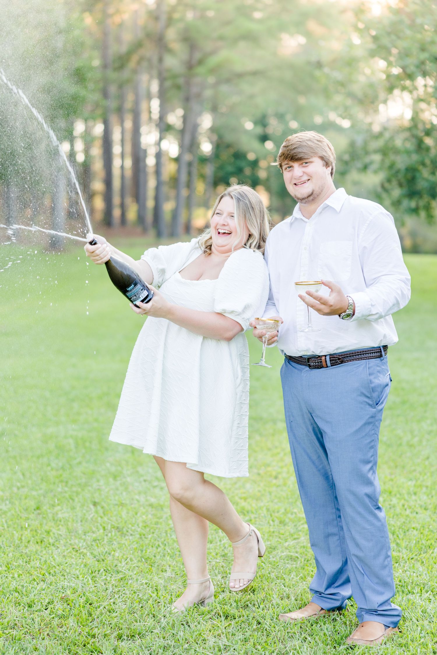 Playful couple popping champagne and celebrating in a park with golden sunlight.