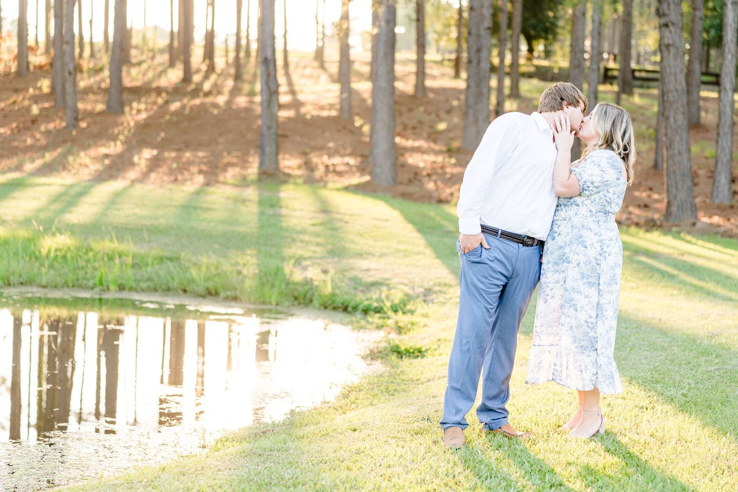 A couple stands near a reflective pond surrounded by pine trees in golden afternoon light.