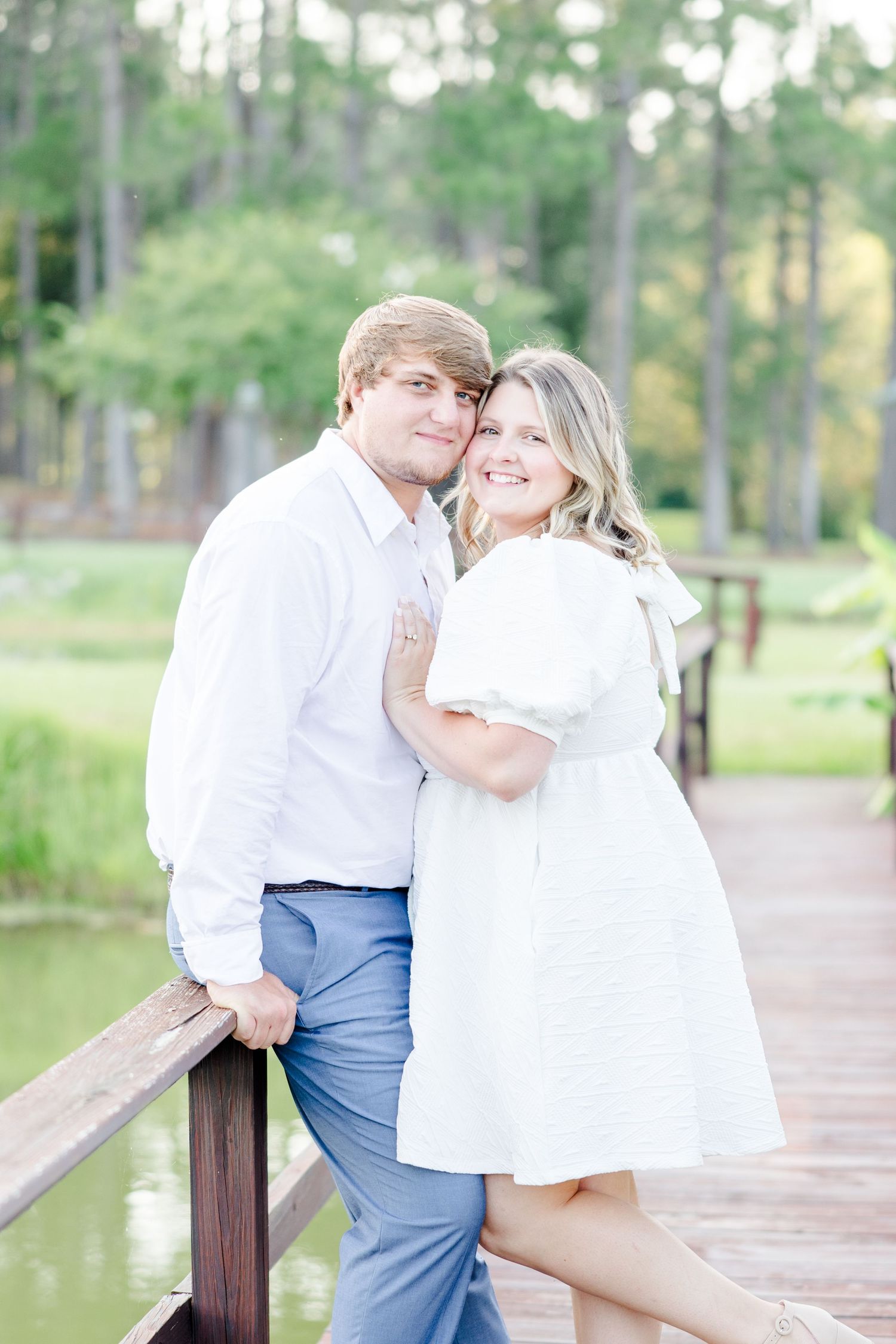 Couple in white clothing sharing romantic moments on a wooden dock surrounded by pine trees.