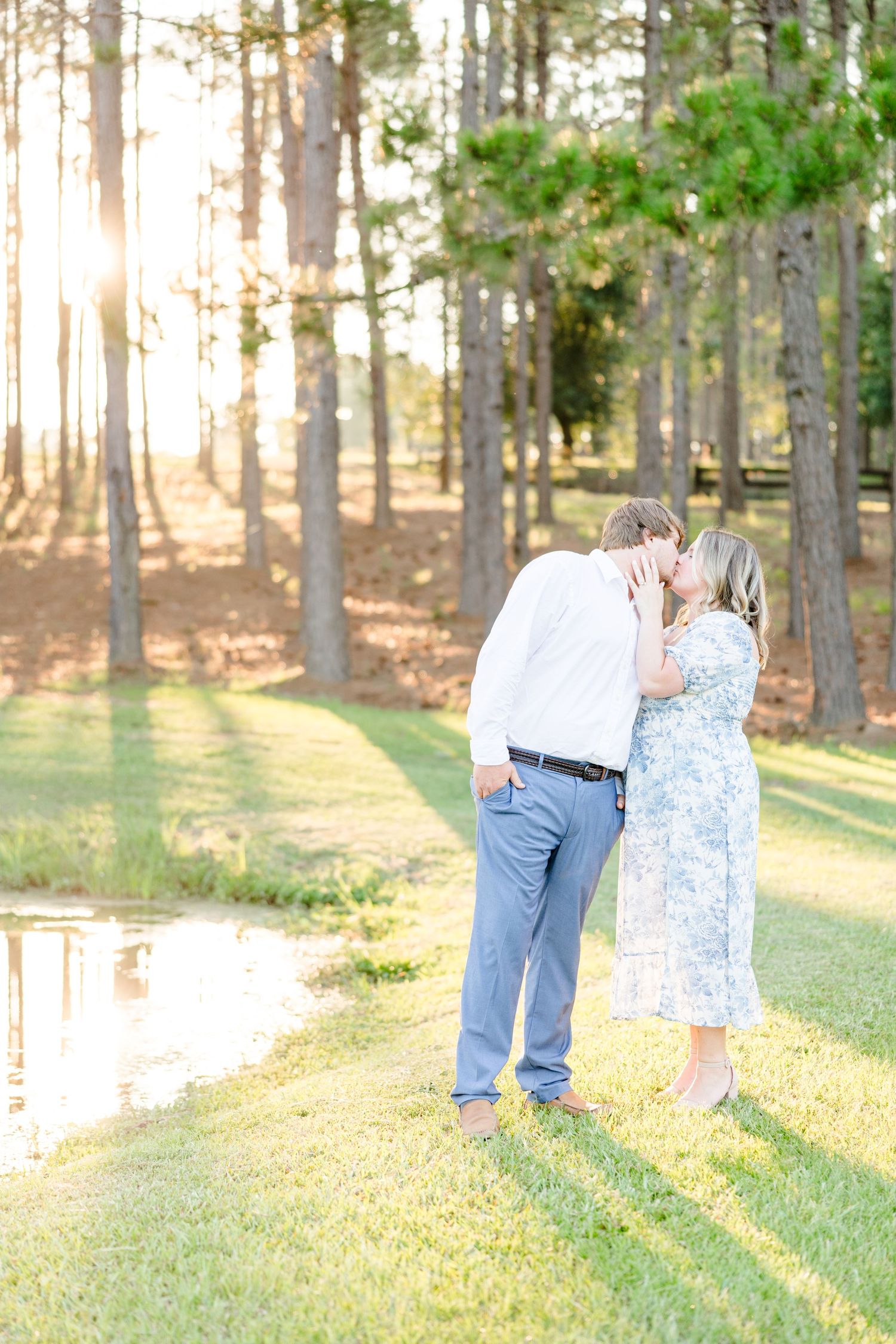 A couple shares a romantic moment by a pond with sunlight filtering through pine trees.