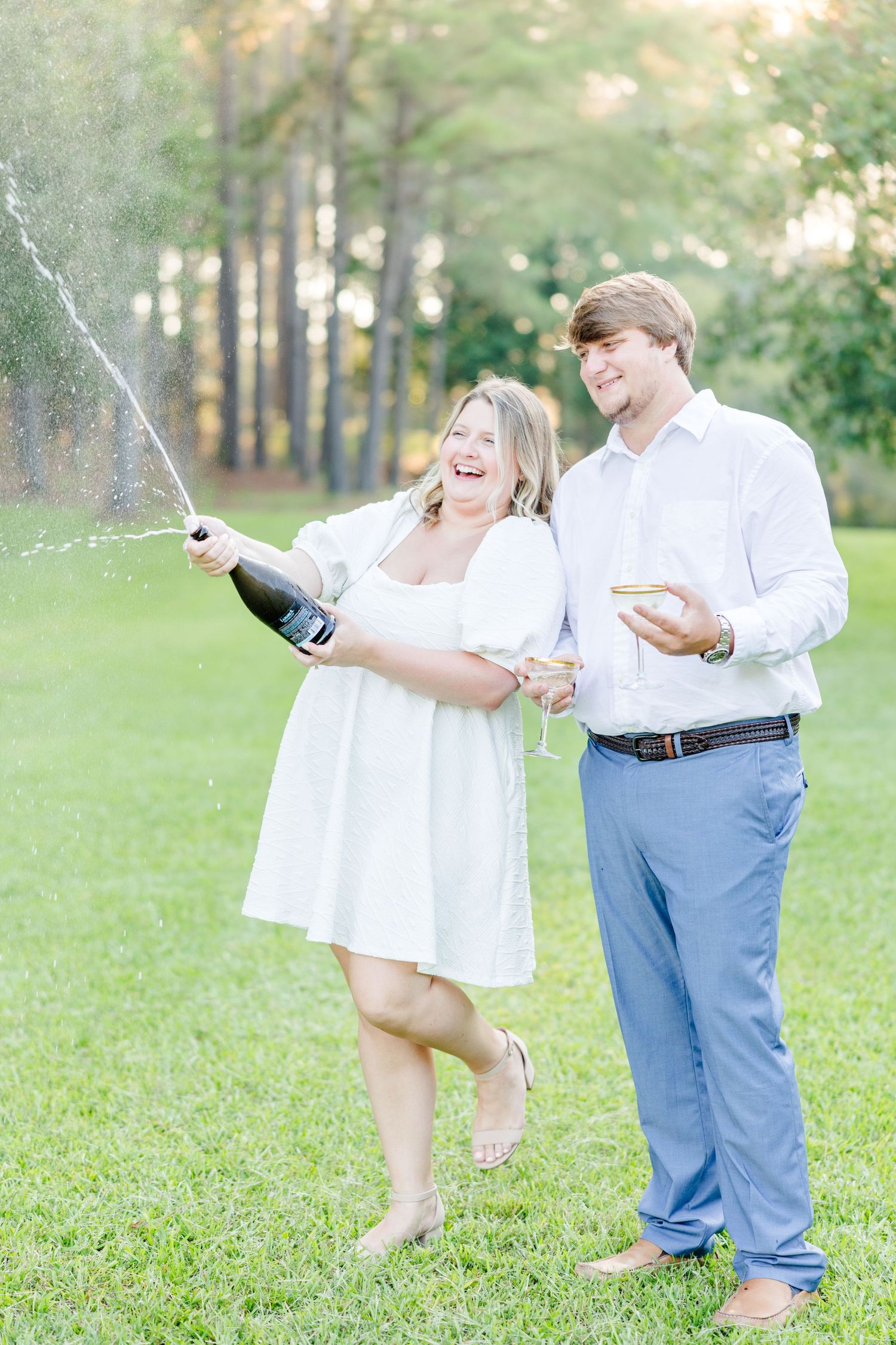 A couple celebrating with champagne in a sunny outdoor setting, spraying and laughing together.