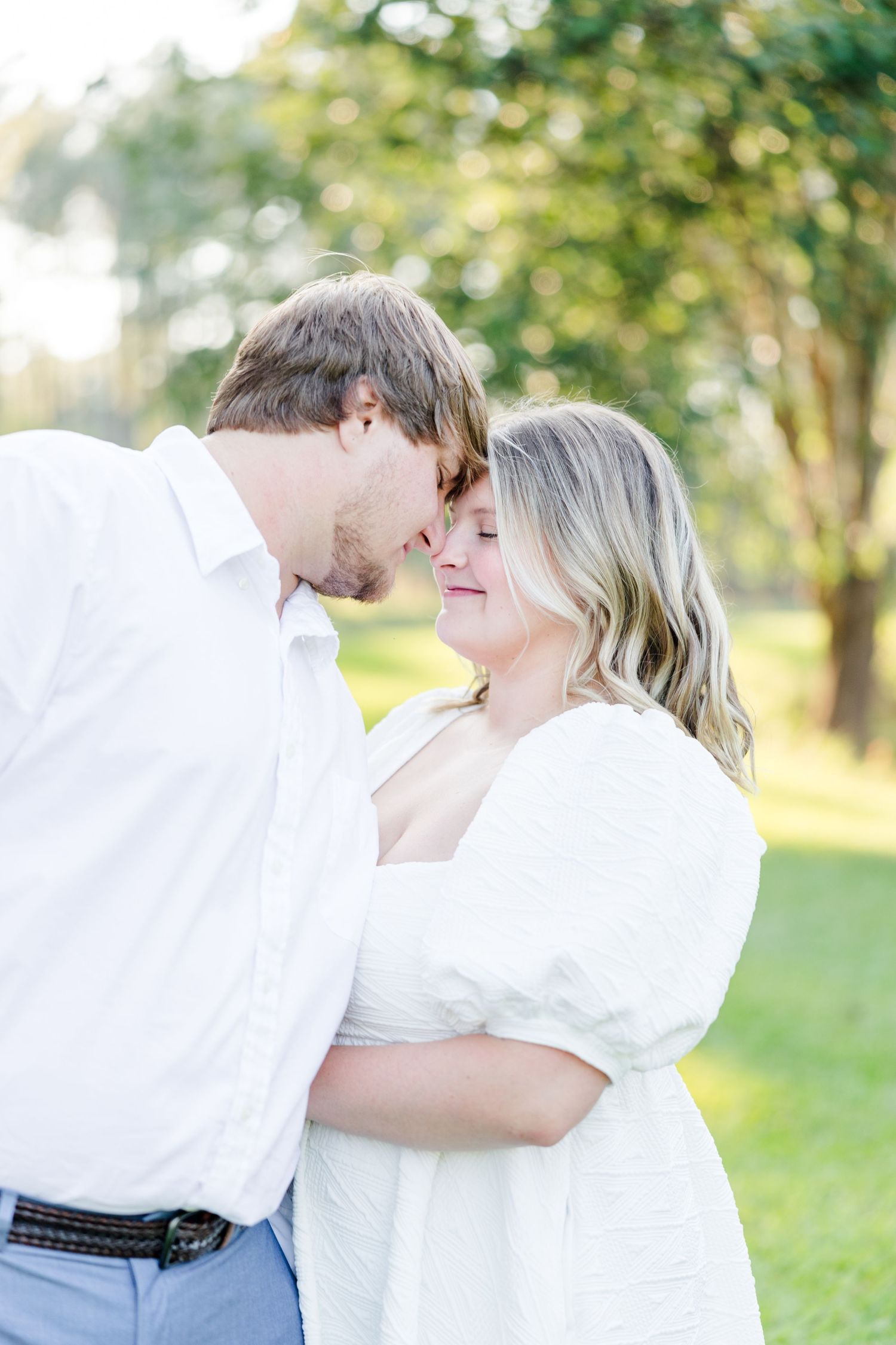 A romantic outdoor engagement photo session with a couple in white outfits embracing in a sunlit park setting.