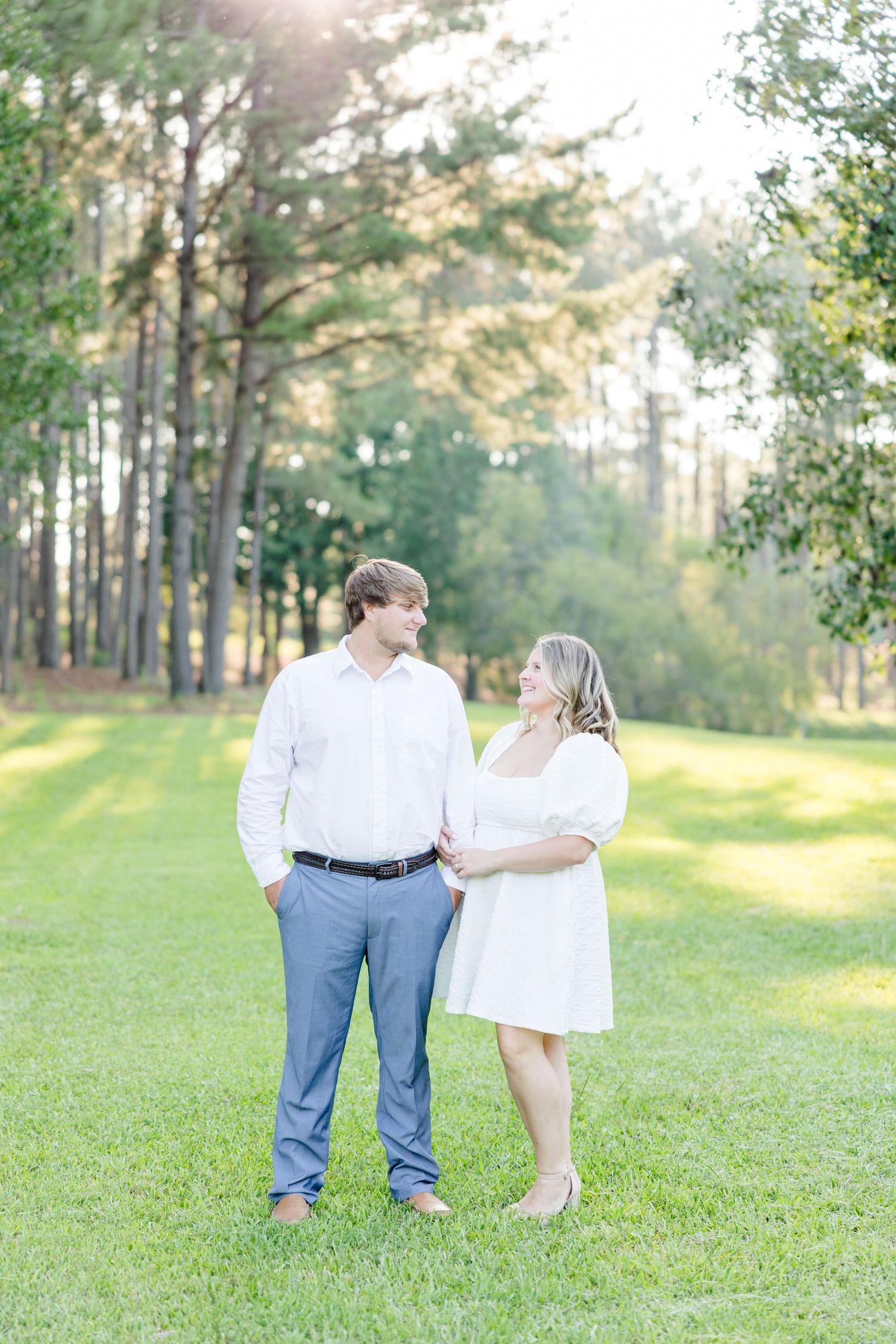 Full length engagement photo of couple standing together on grassy lawn with pine trees.