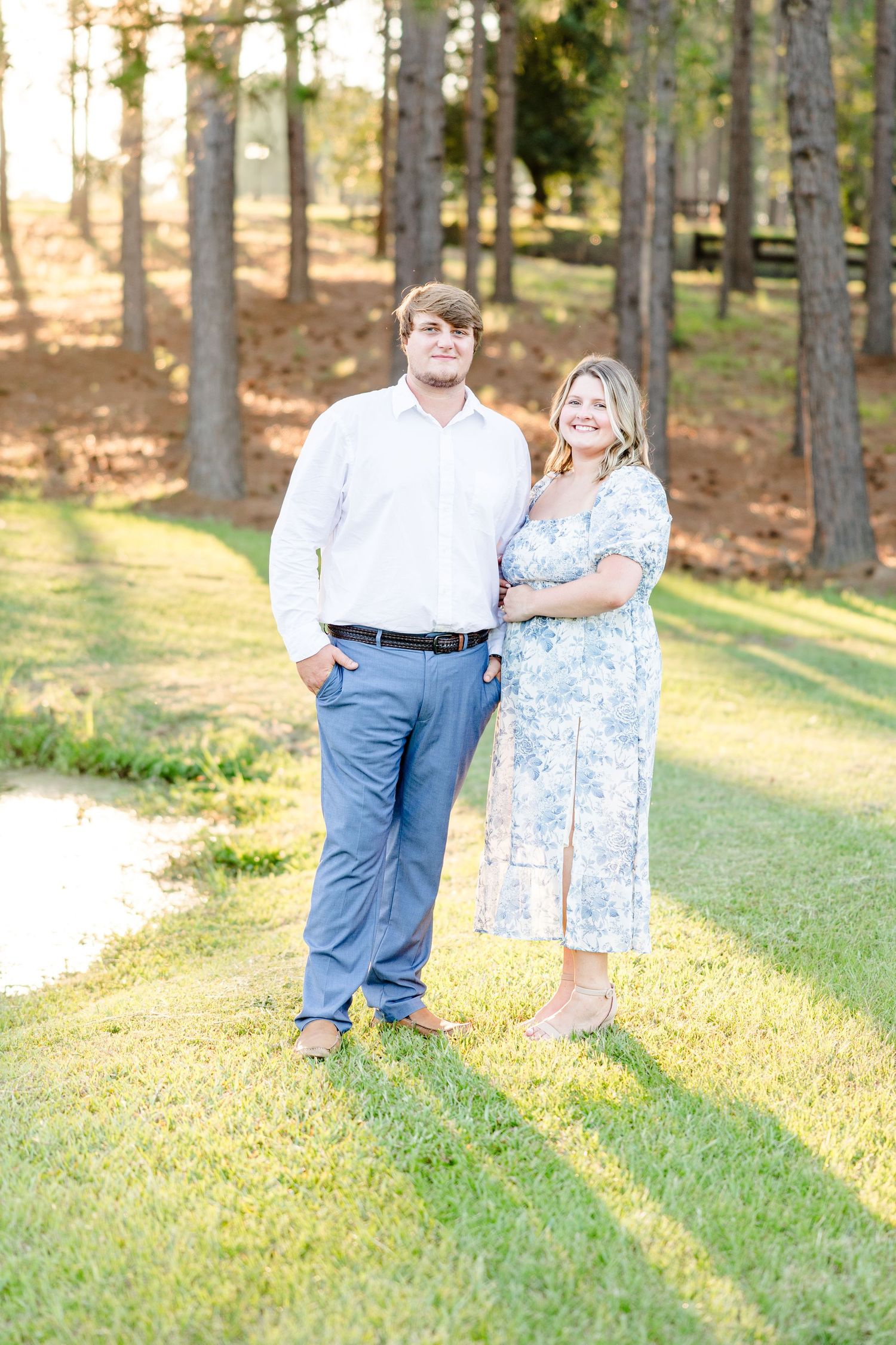 A couple stands together on a grassy hill with pine trees in the background during sunset.