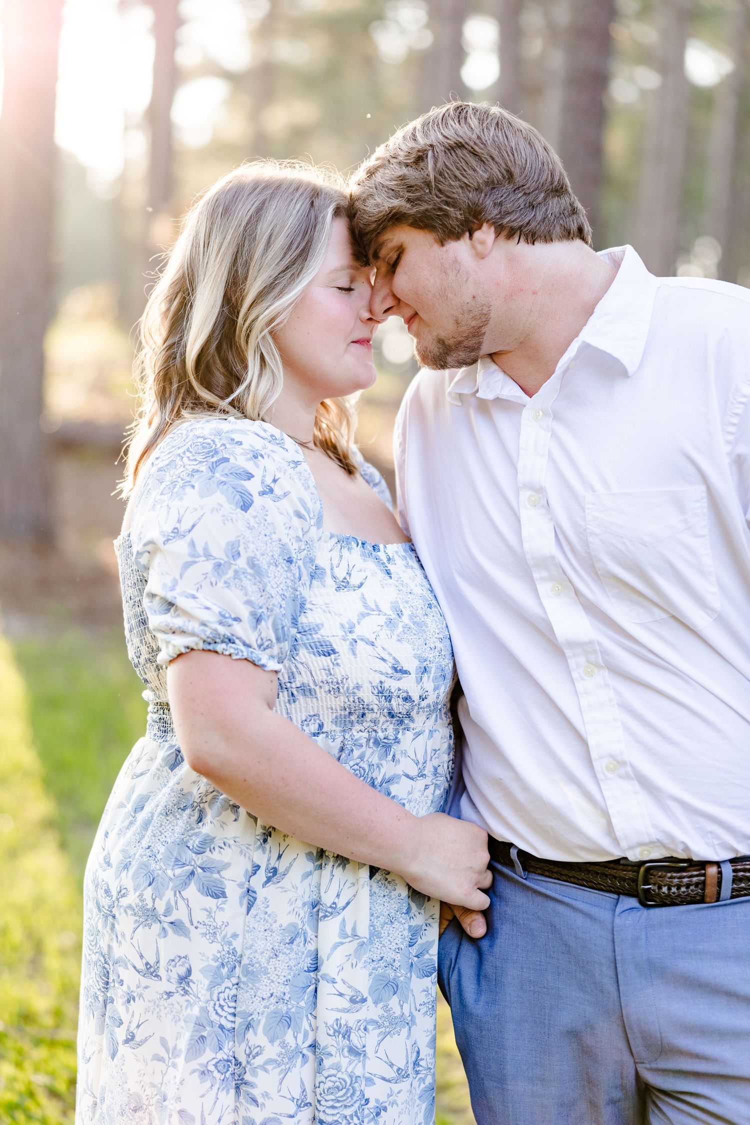 A couple shares an intimate moment in a sunlit forest wearing blue and white attire.