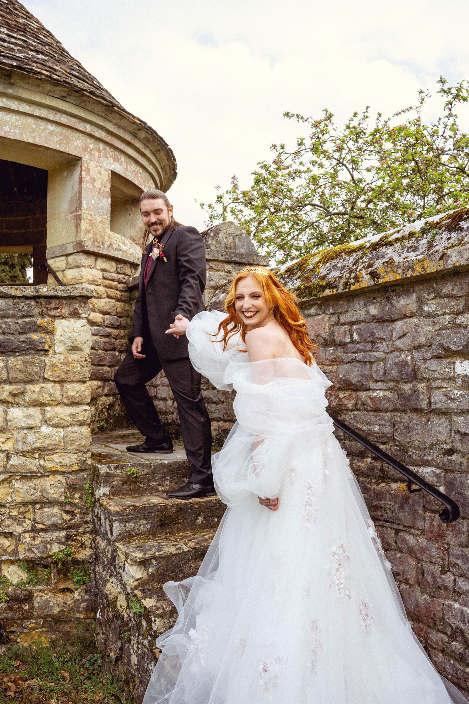 Joyful bride in embellished white gown, hand-in-hand with groom in black suit, laughing on old brick staircase in the So