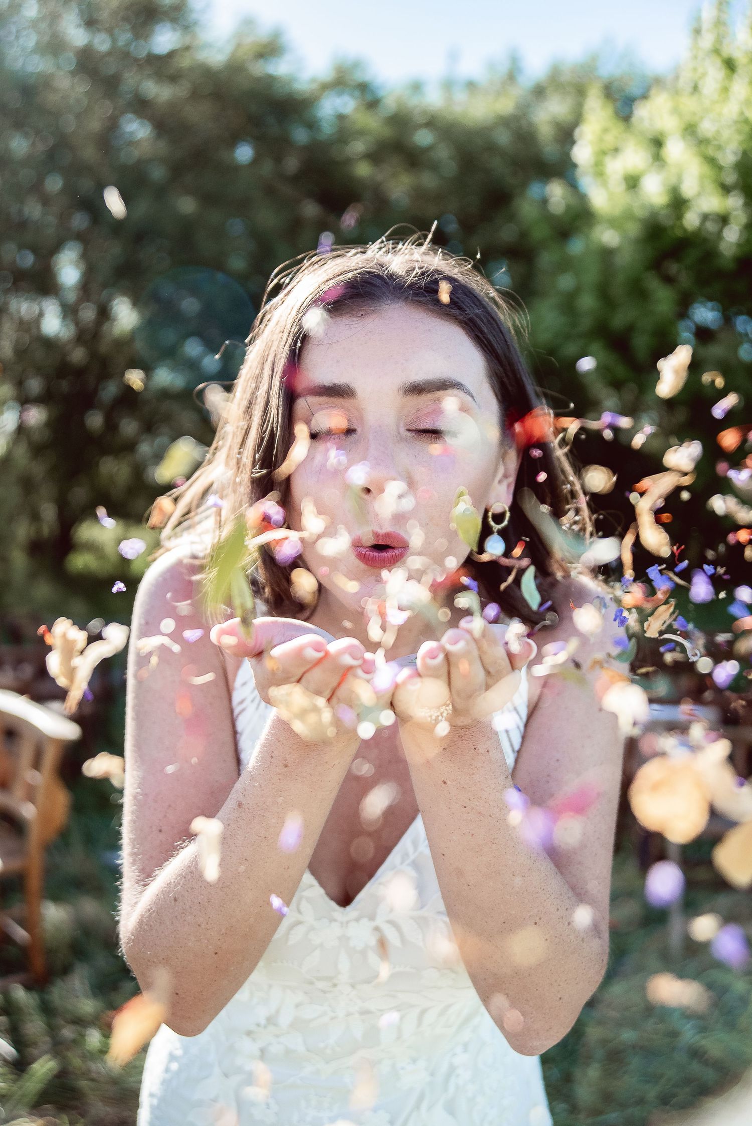 Young bride in sunlit outdoor scene, blowing colourful confetti in celebration, adorned in a sleek white dress with flor