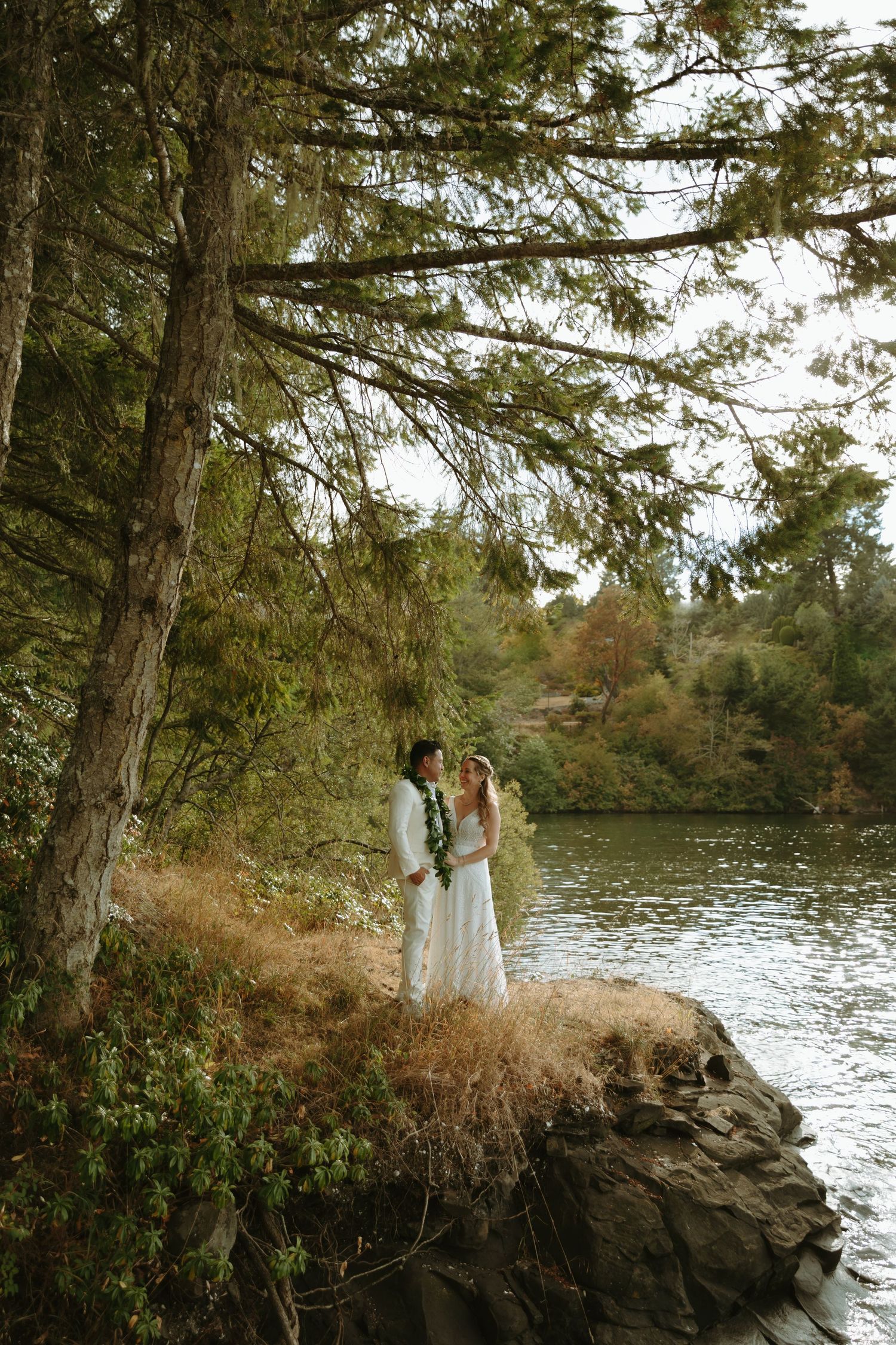 A romantic wedding portrait by a lake with tall evergreen trees in the background.