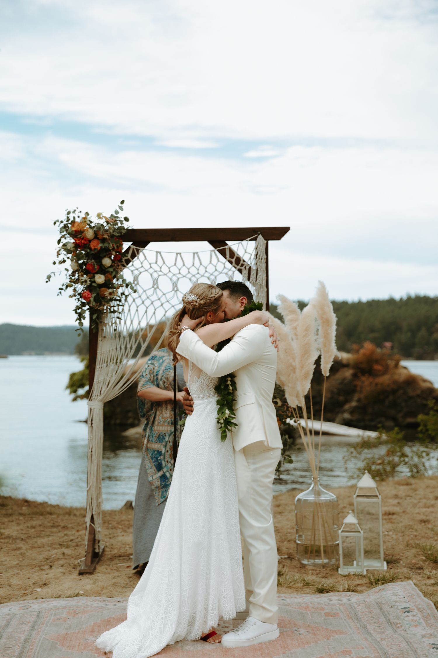 A romantic moment under a macrame wedding arch decorated with orange flowers by the water.