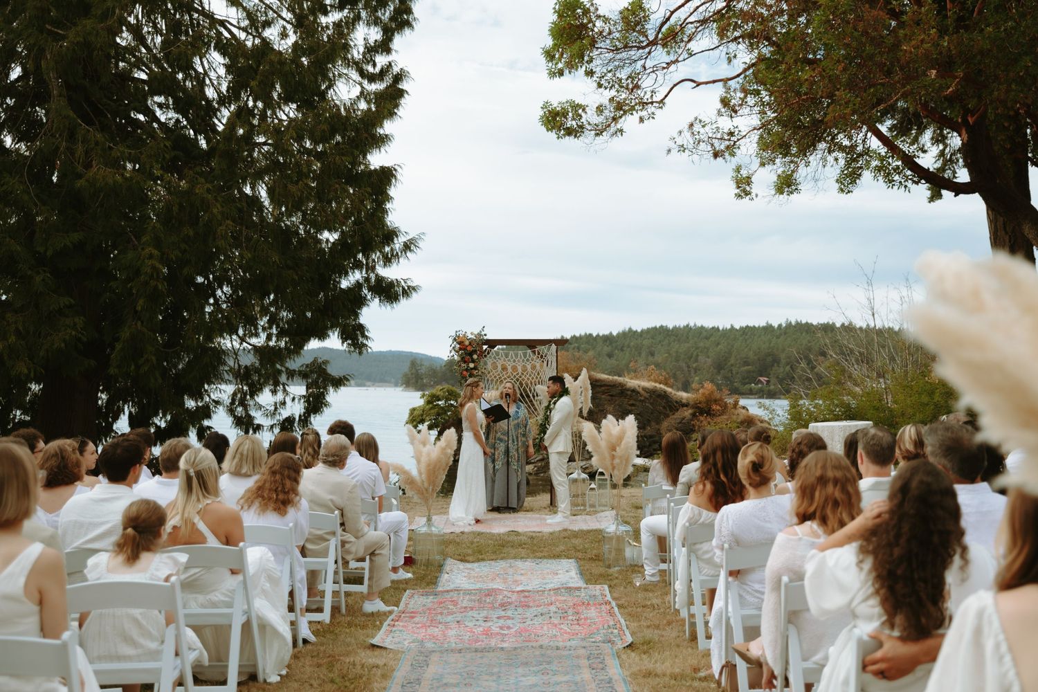 A scenic outdoor wedding ceremony setup with white chairs and a floral arch overlooking a lake.