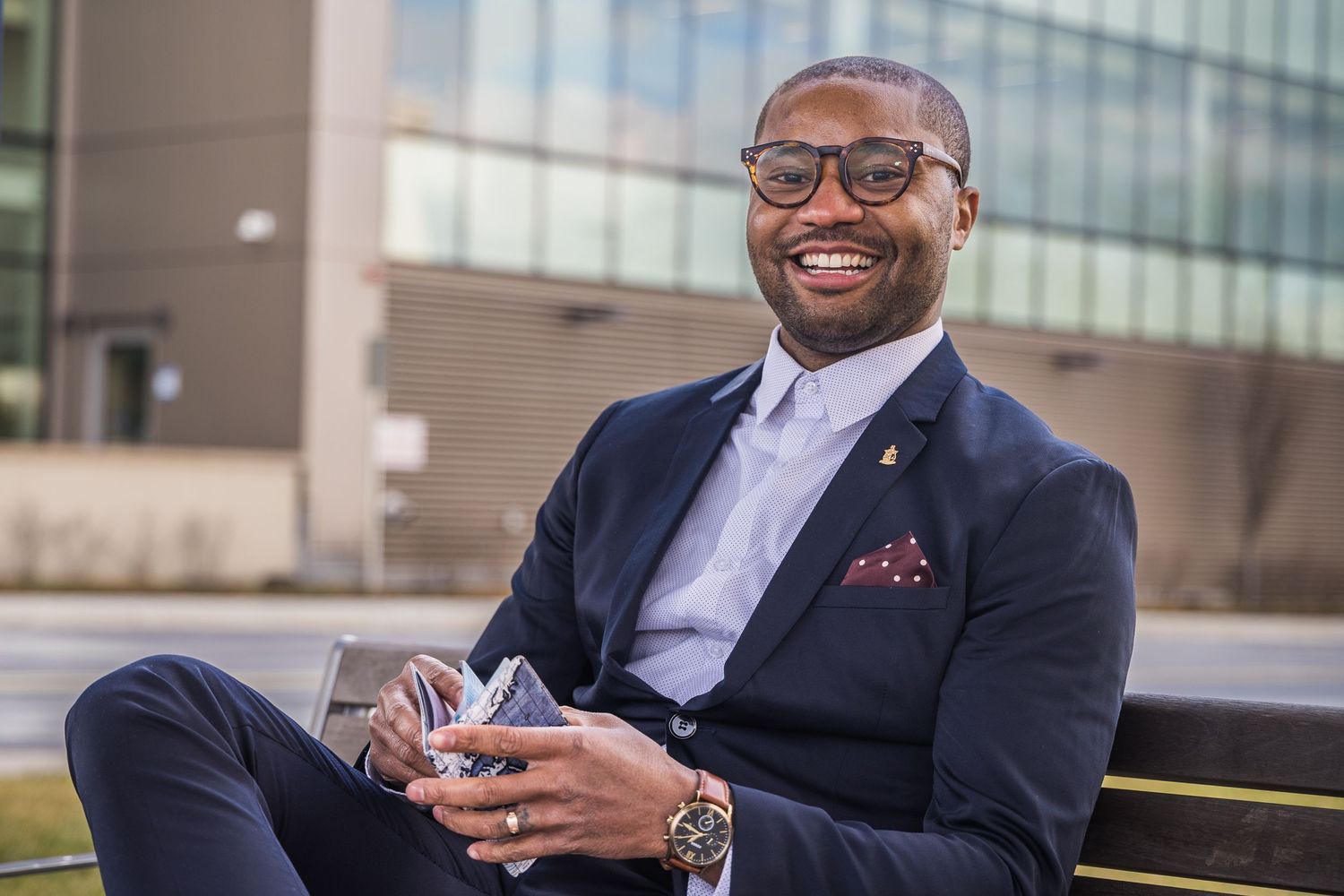Professional in navy suit and glasses sits outdoors with mobile phone near modern building.