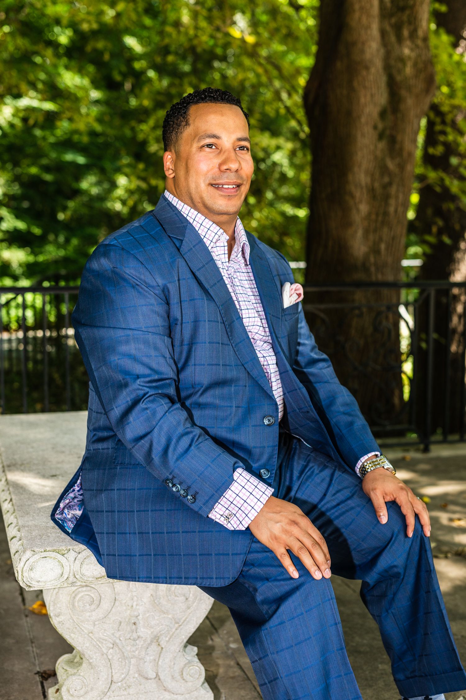 Businessman in blue plaid suit and pink striped shirt sits confidently on stone bench outdoors.