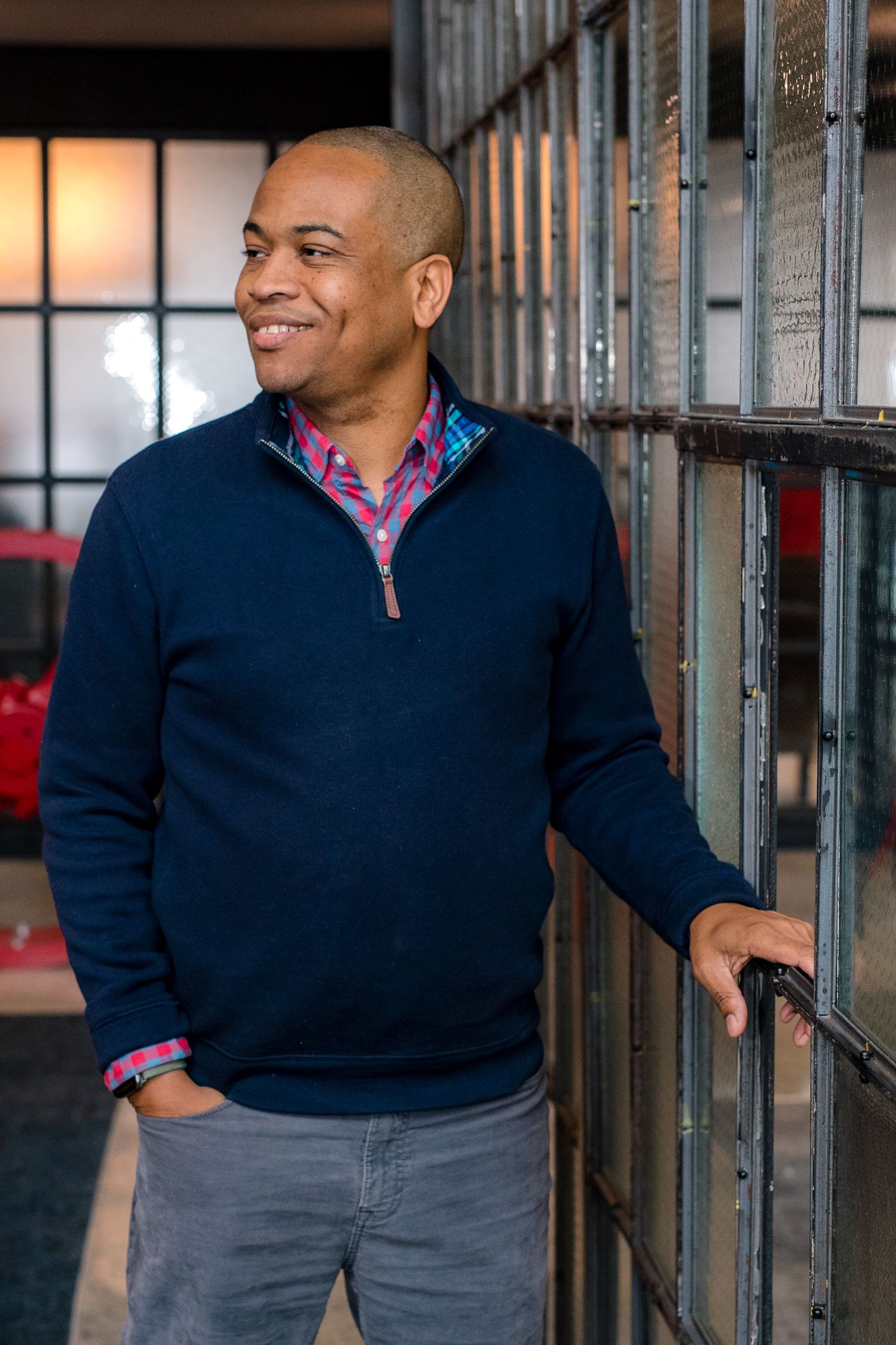 Professional wearing a navy quarter-zip sweater and pink checkered shirt poses casually against an industrial backdrop.