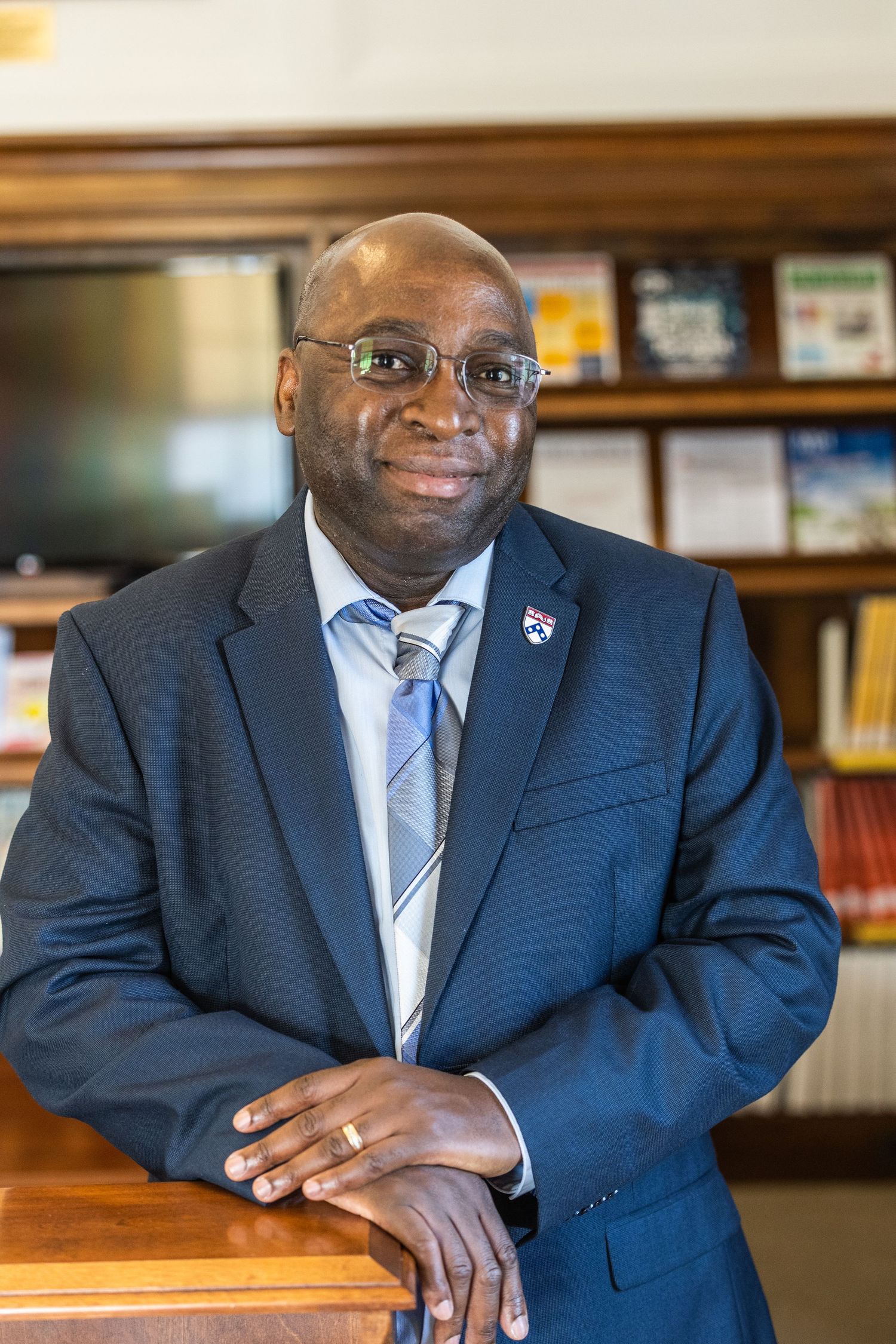 Professional in navy suit and tie stands confidently in office with bookshelf background.