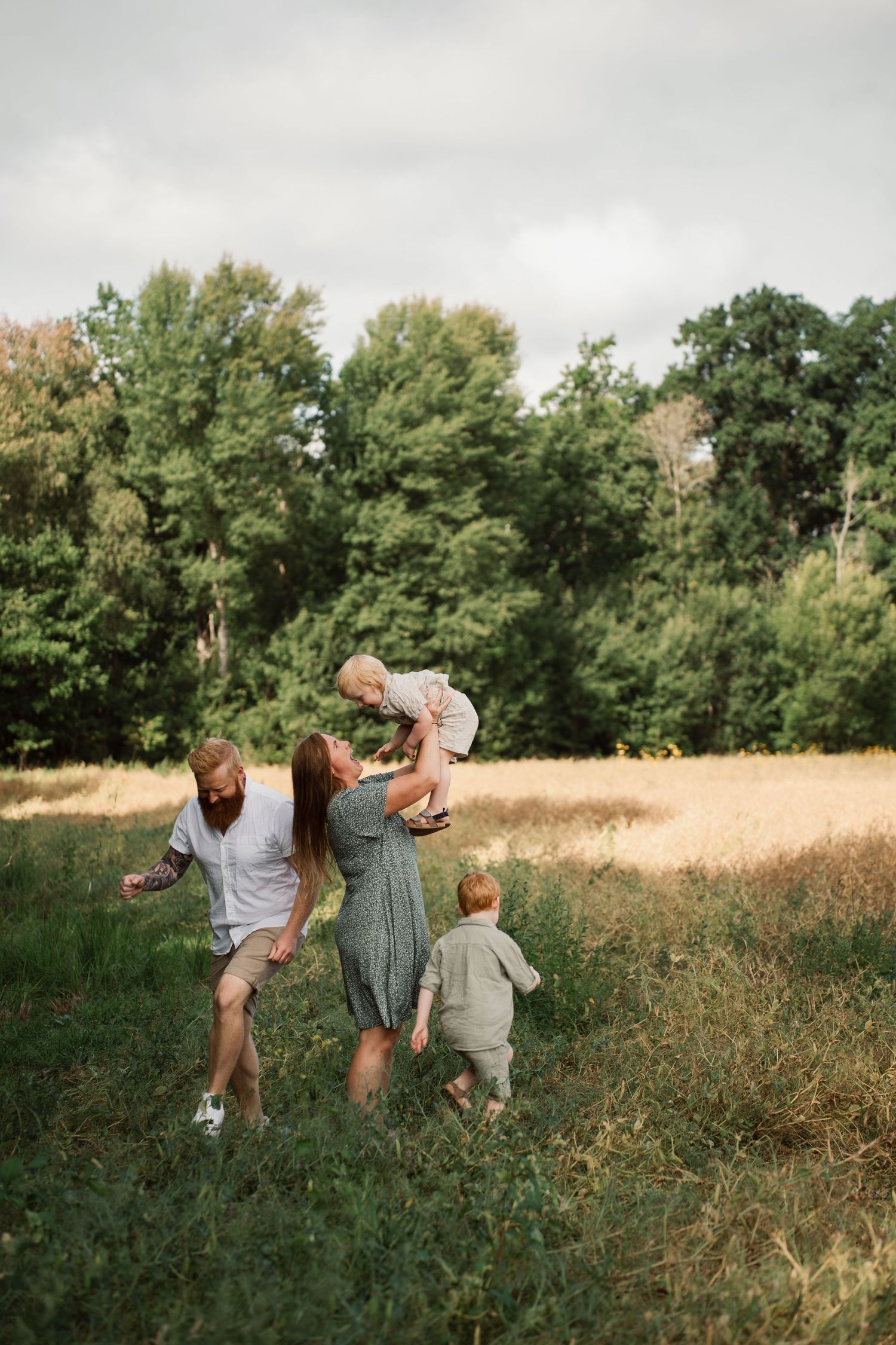 A family runs playfully through a sunlit meadow surrounded by tall green trees.