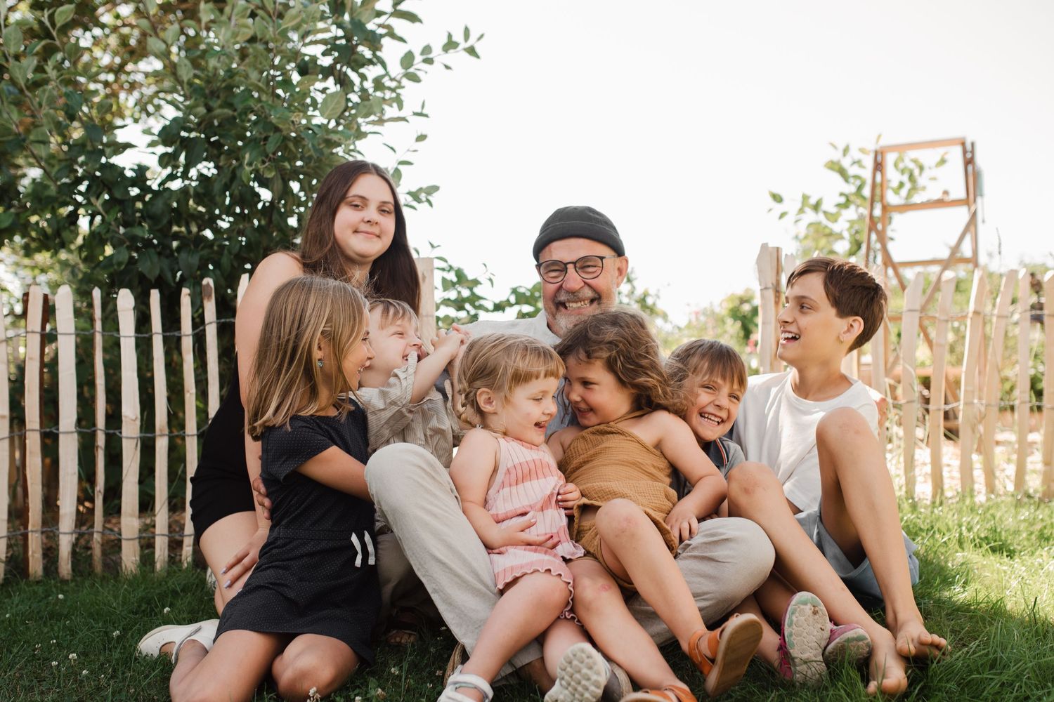 A family group sits together on grass near a rustic wooden fence.