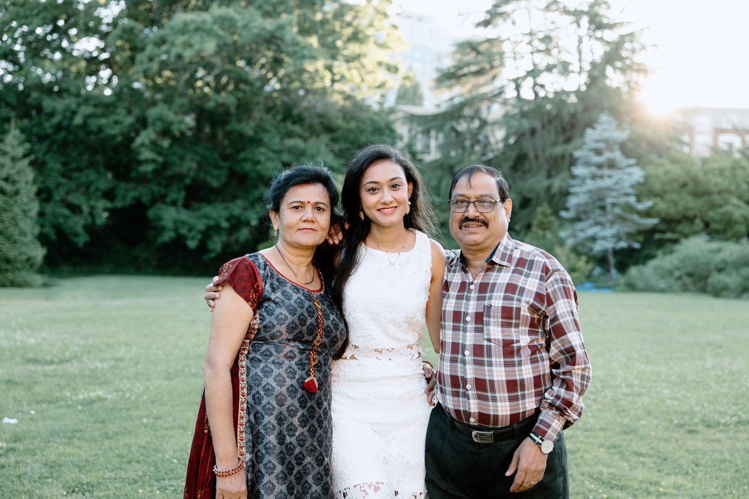Family portrait outdoors with daughter in white dress and parents in casual attire.