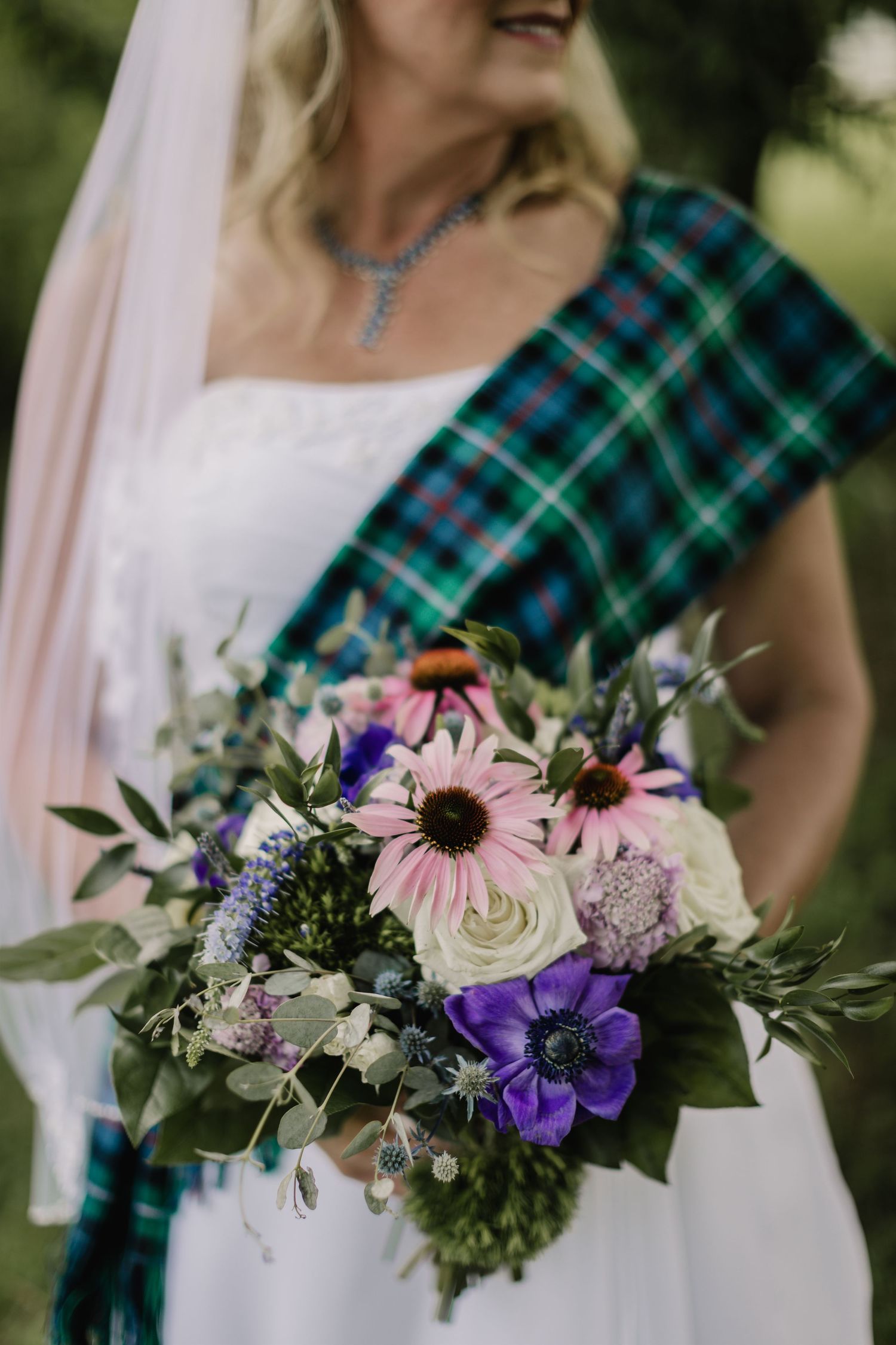 A bridal bouquet with pink, purple and white flowers against a tartan sash and white dress.