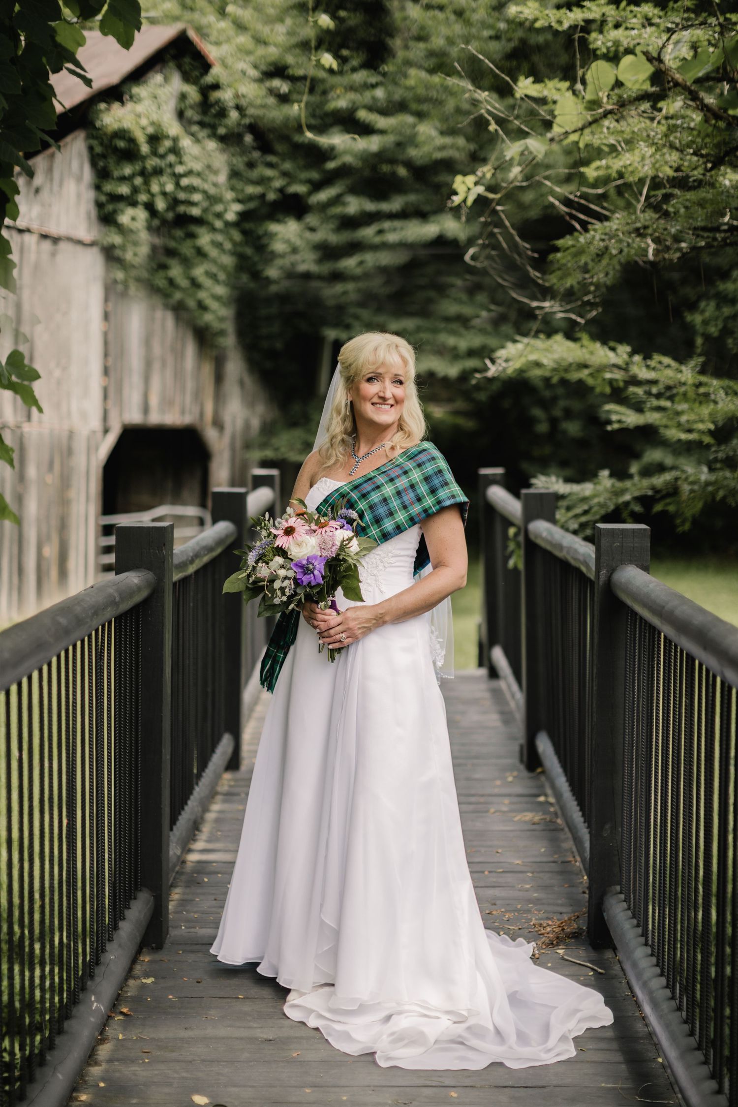 Bride in elegant white gown poses on wooden bridge with Scottish tartan accent.
