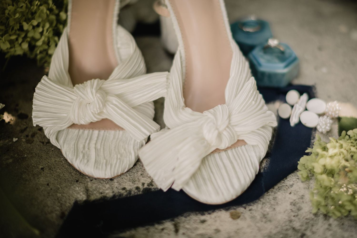 Elegant white bridal shoes with pleated bow detail on concrete surface.