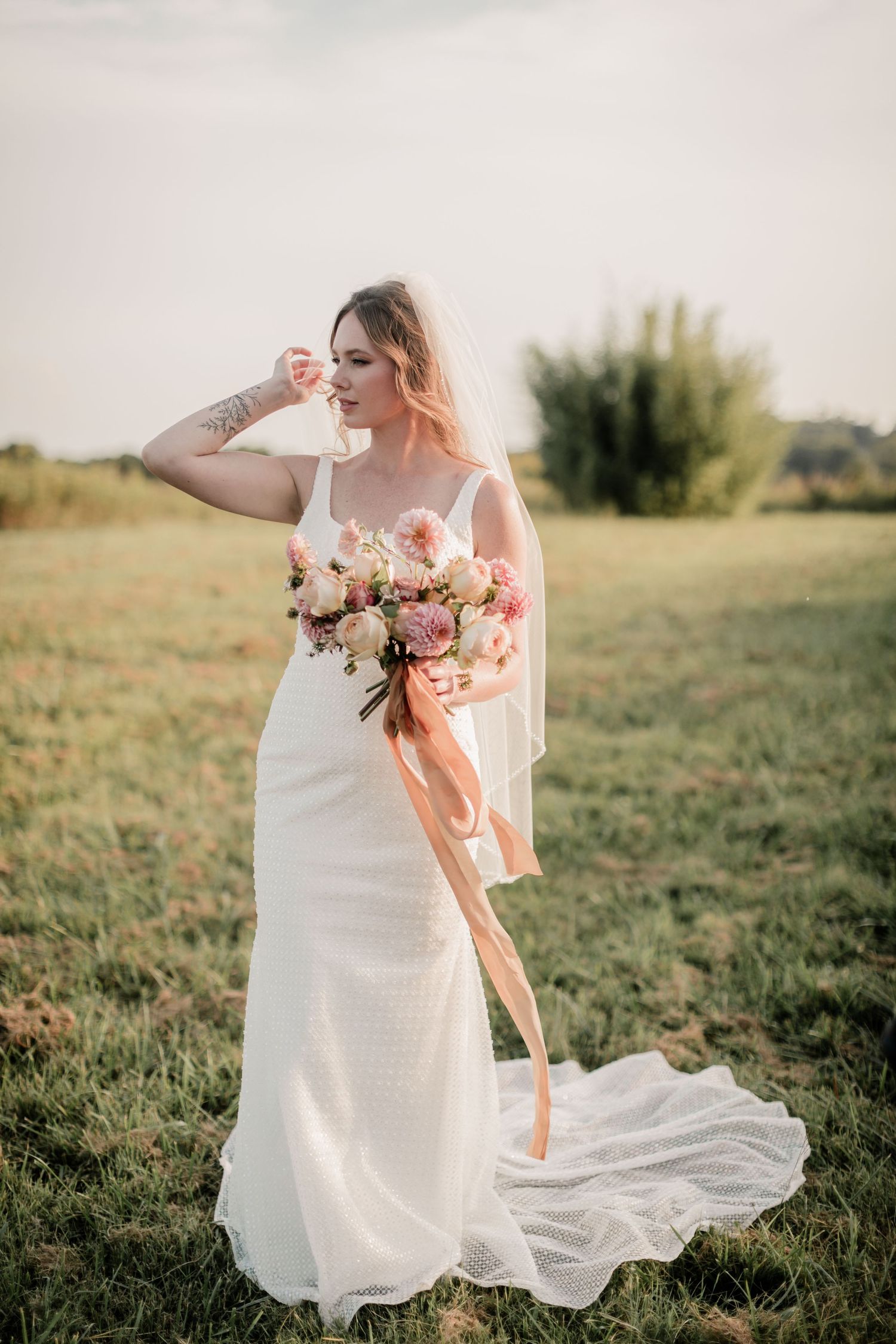 A bride in a flowing white dress holds a peach and pink bouquet while standing in a sunlit field.