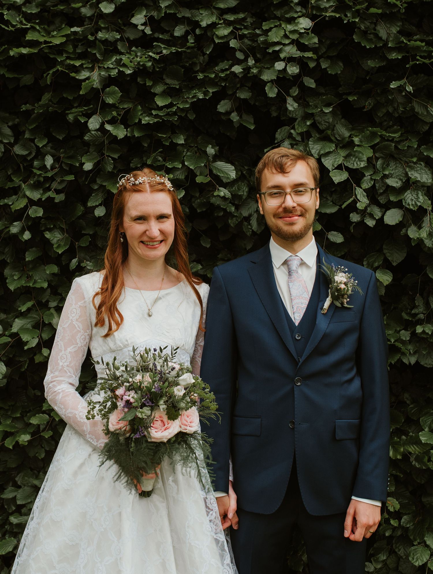 Wedding couple standing against lush green hedge backdrop with floral bouquet.