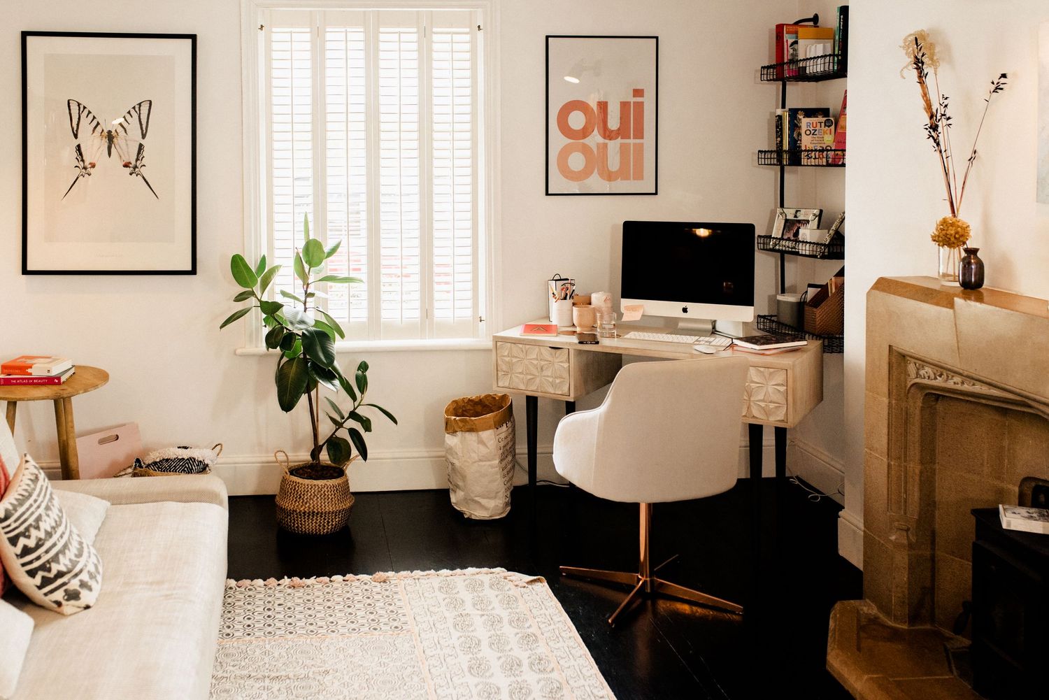 Minimalist home office with butterfly art, wooden desk, and white chair against window with shutters.