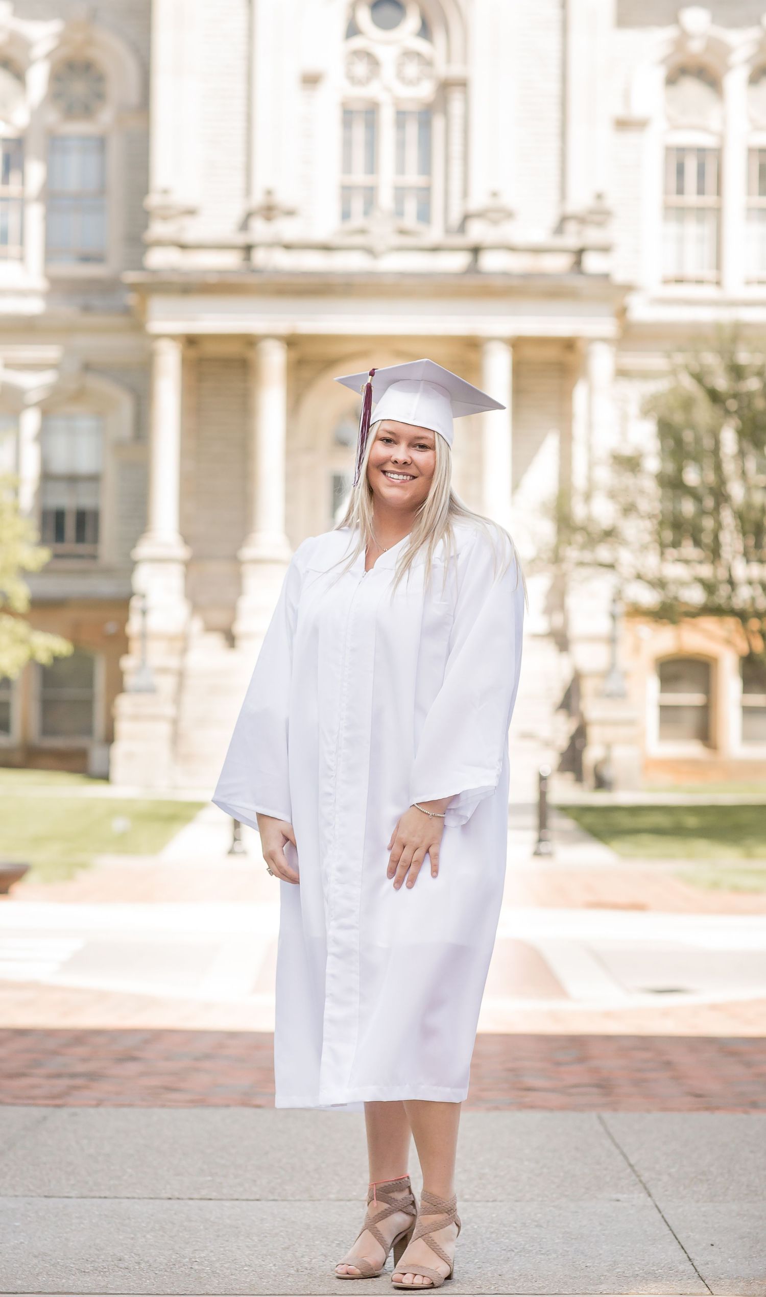 Graduate in white cap and gown smiling in front of a grand historic building outdoors.