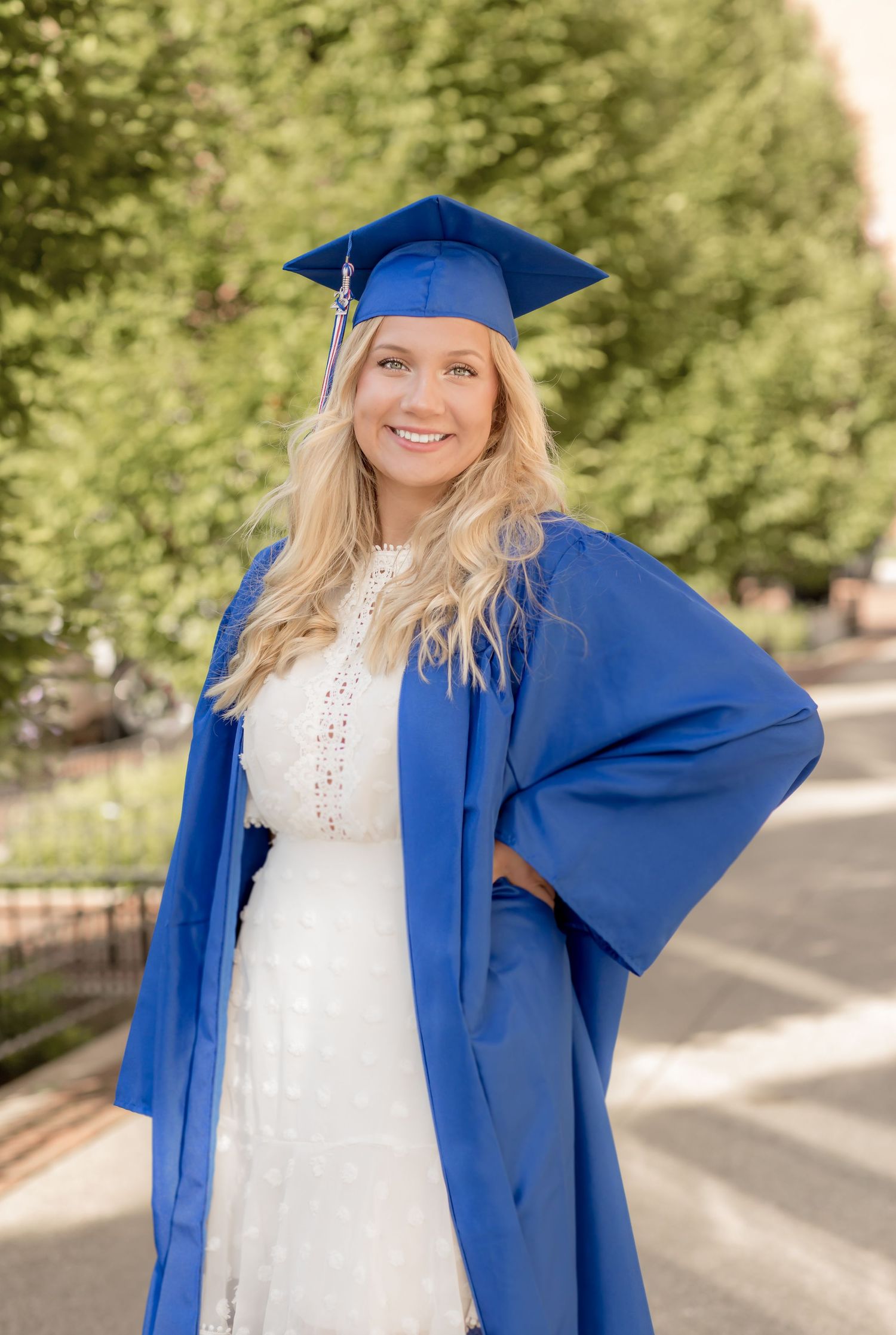 Smiling blonde graduate standing in blue cap and gown on a tree-lined campus walkway.