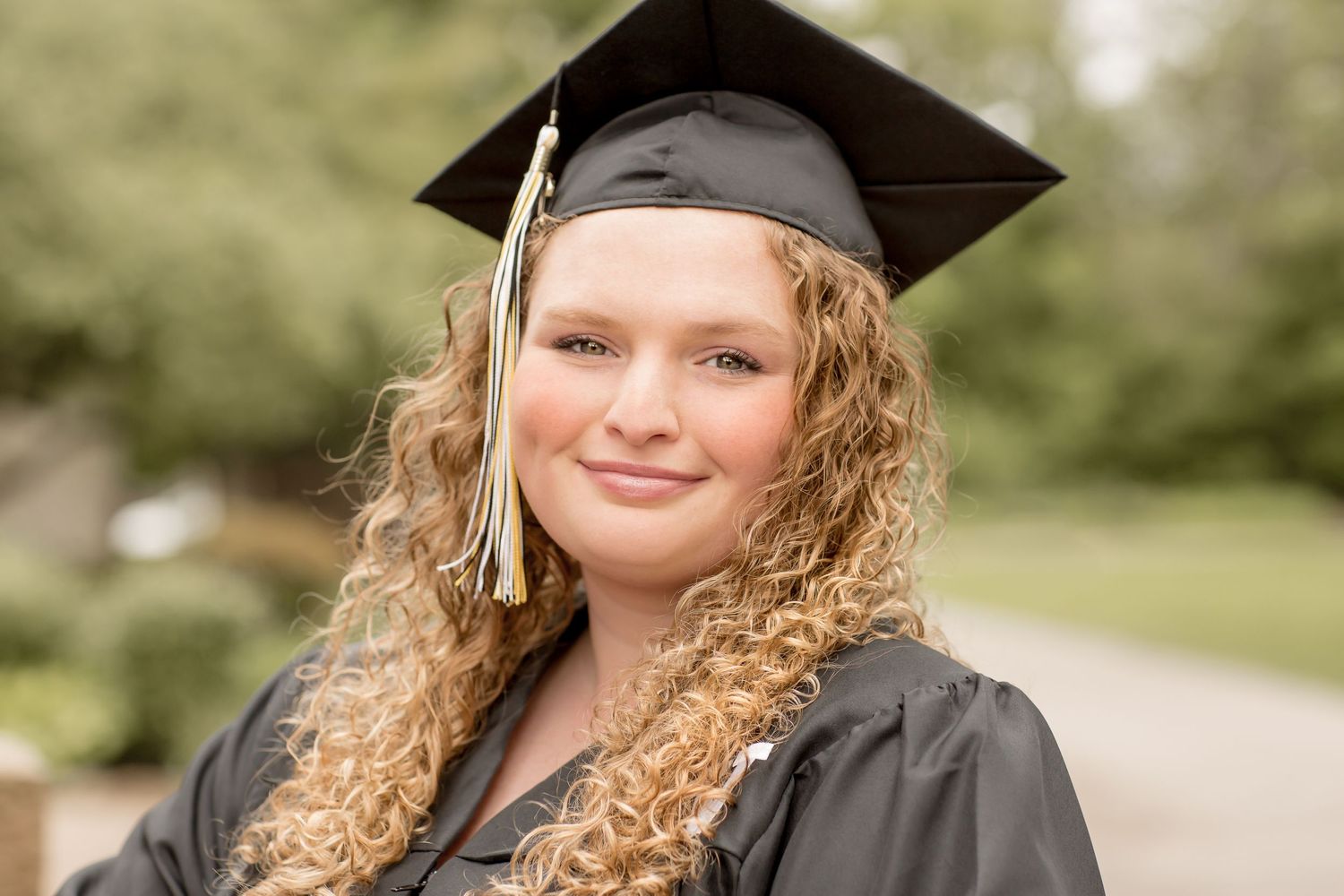Curly-haired graduate smiles in black cap and gown outdoors.
