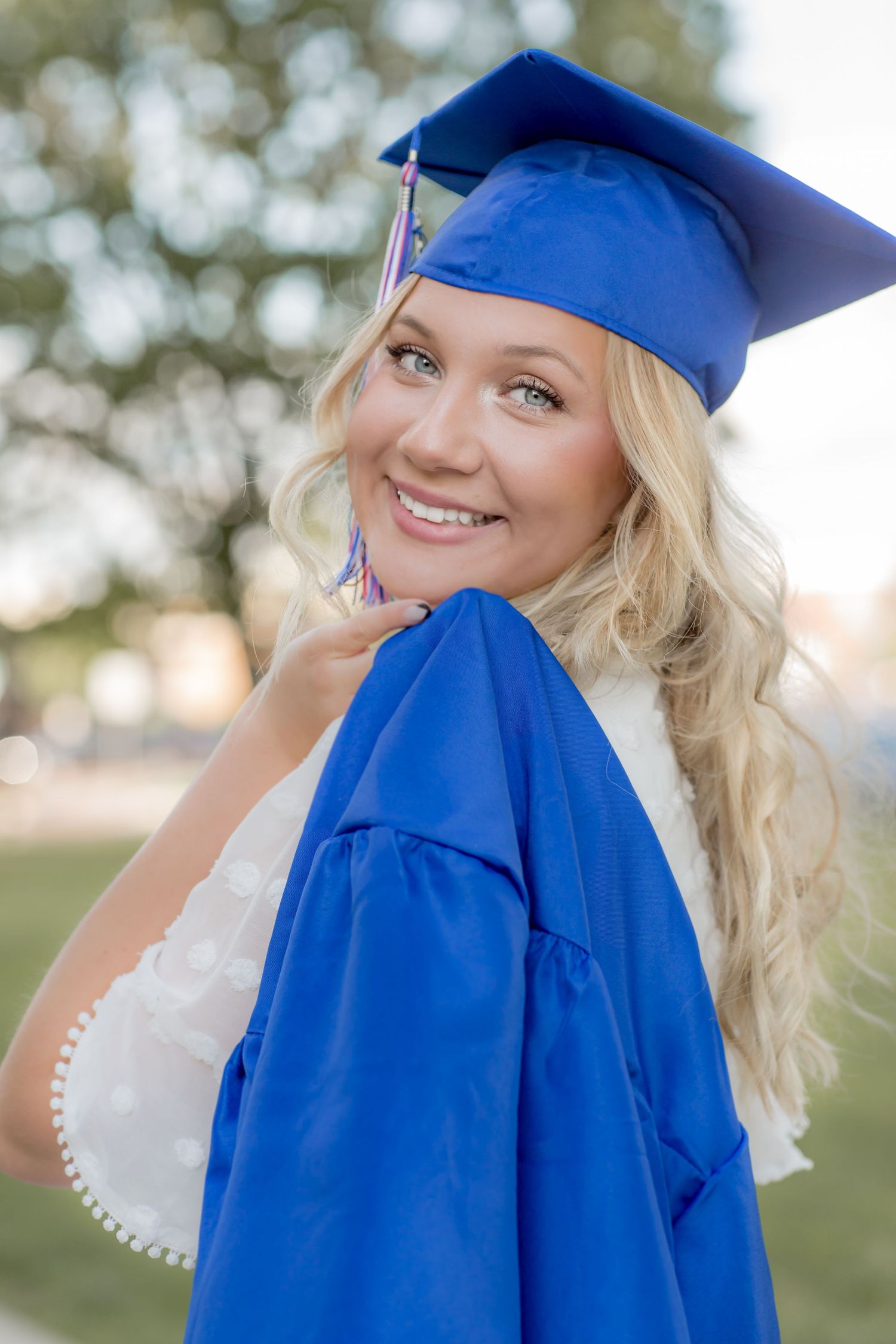 Blonde graduate in blue cap and gown smiling over her shoulder in an outdoor setting.