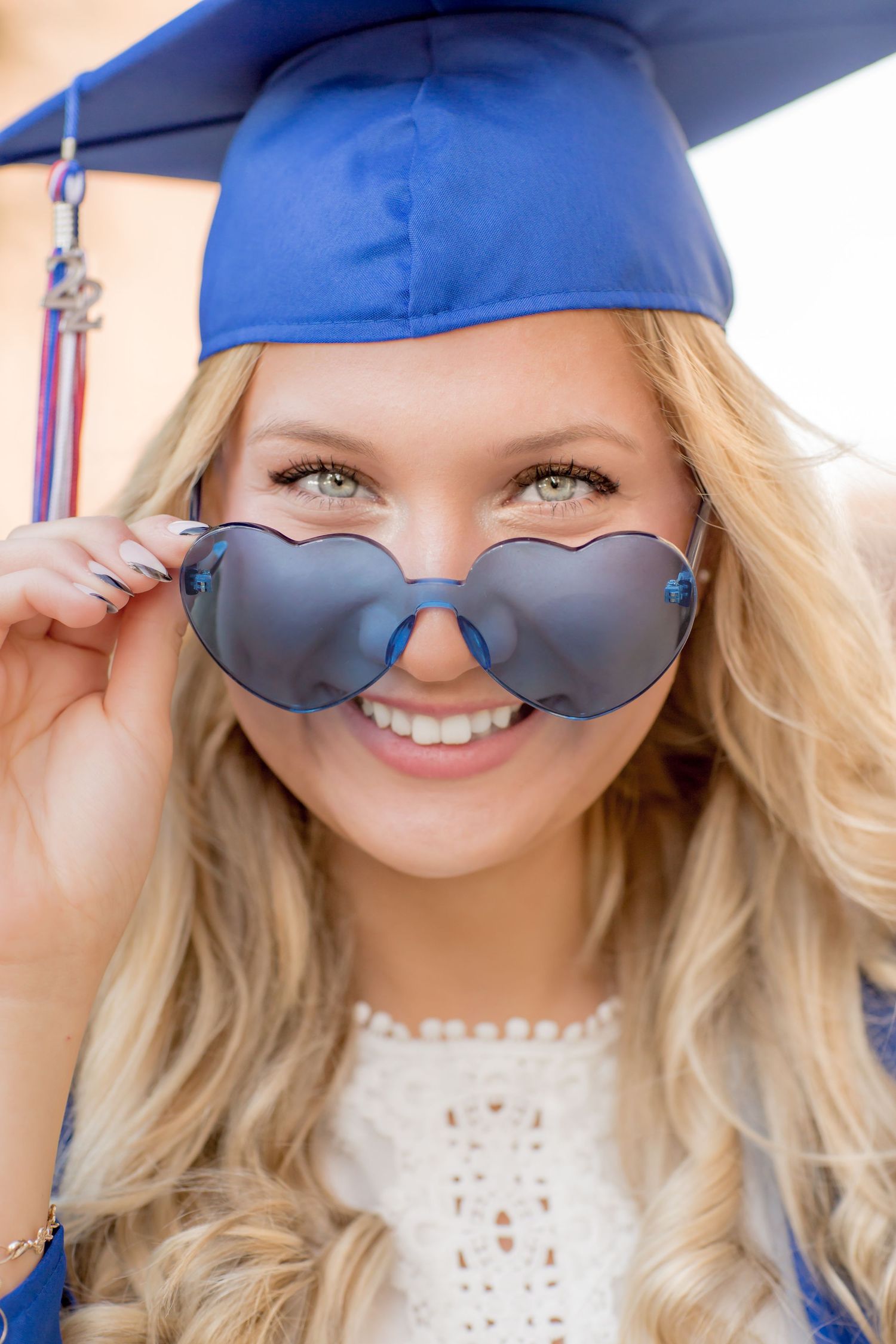 Blonde graduate in blue cap holds heart-shaped sunglasses outdoors.