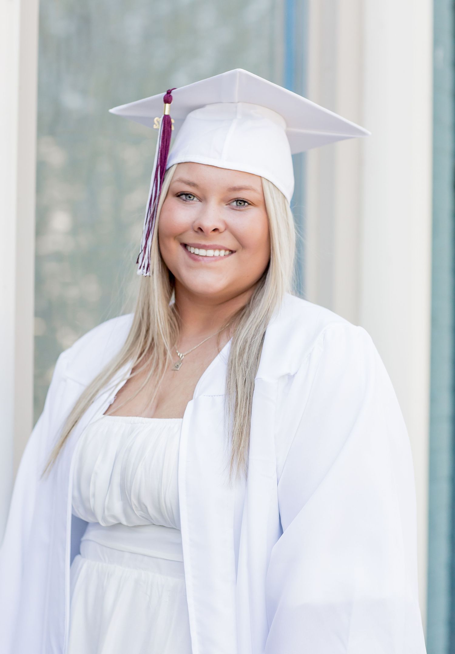 Smiling blonde graduate in white cap and gown posing near bright windows in natural light.