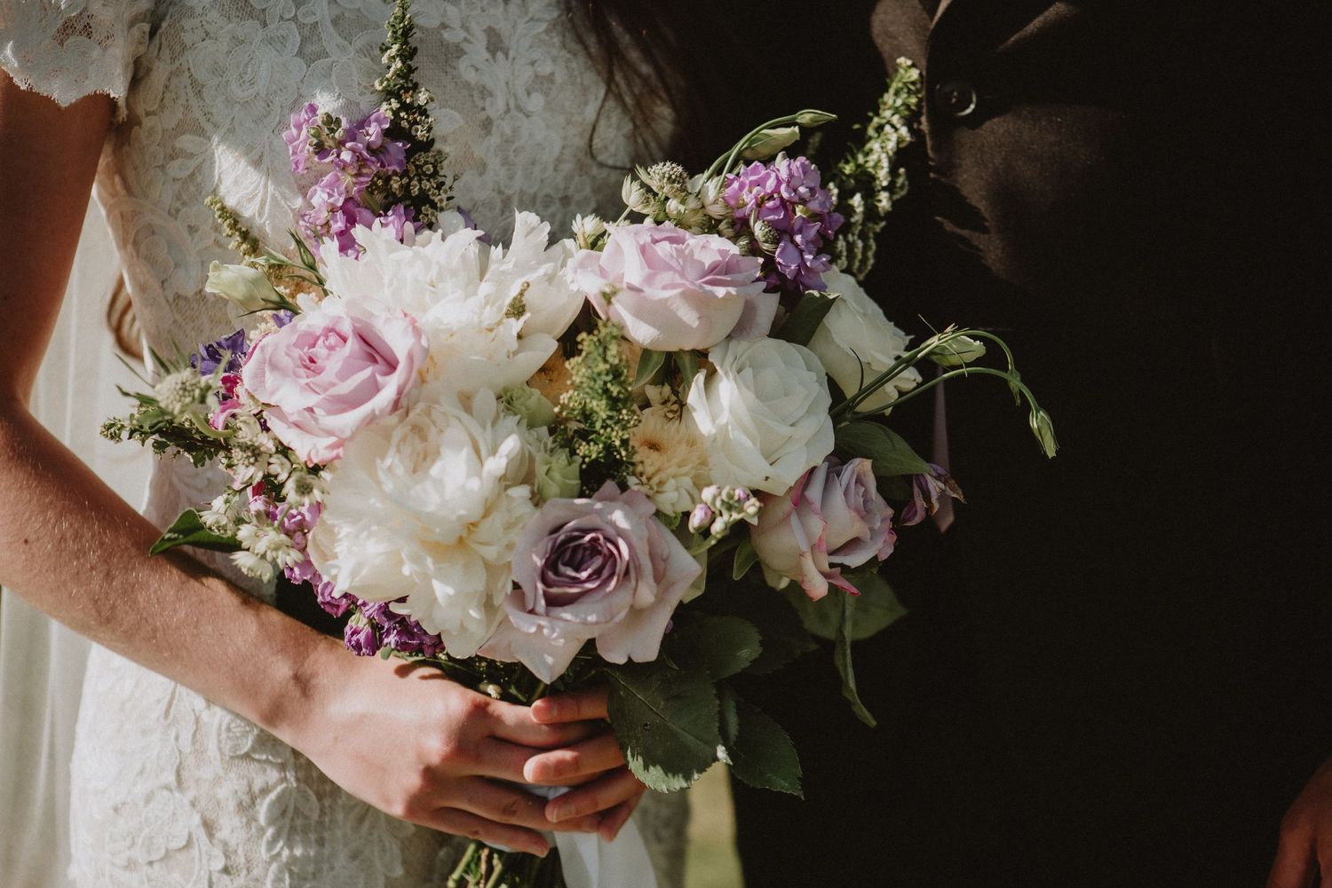 A delicate wedding bouquet featuring purple roses, white peonies and greenery held against a dark background.