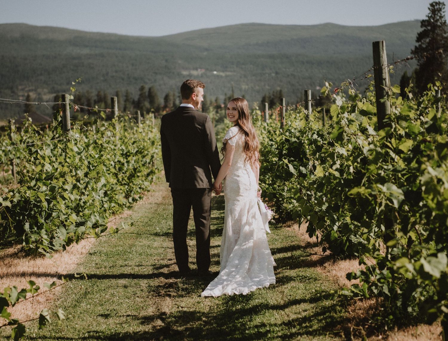 Couple walking hand in hand through vineyard rows in wedding attire.