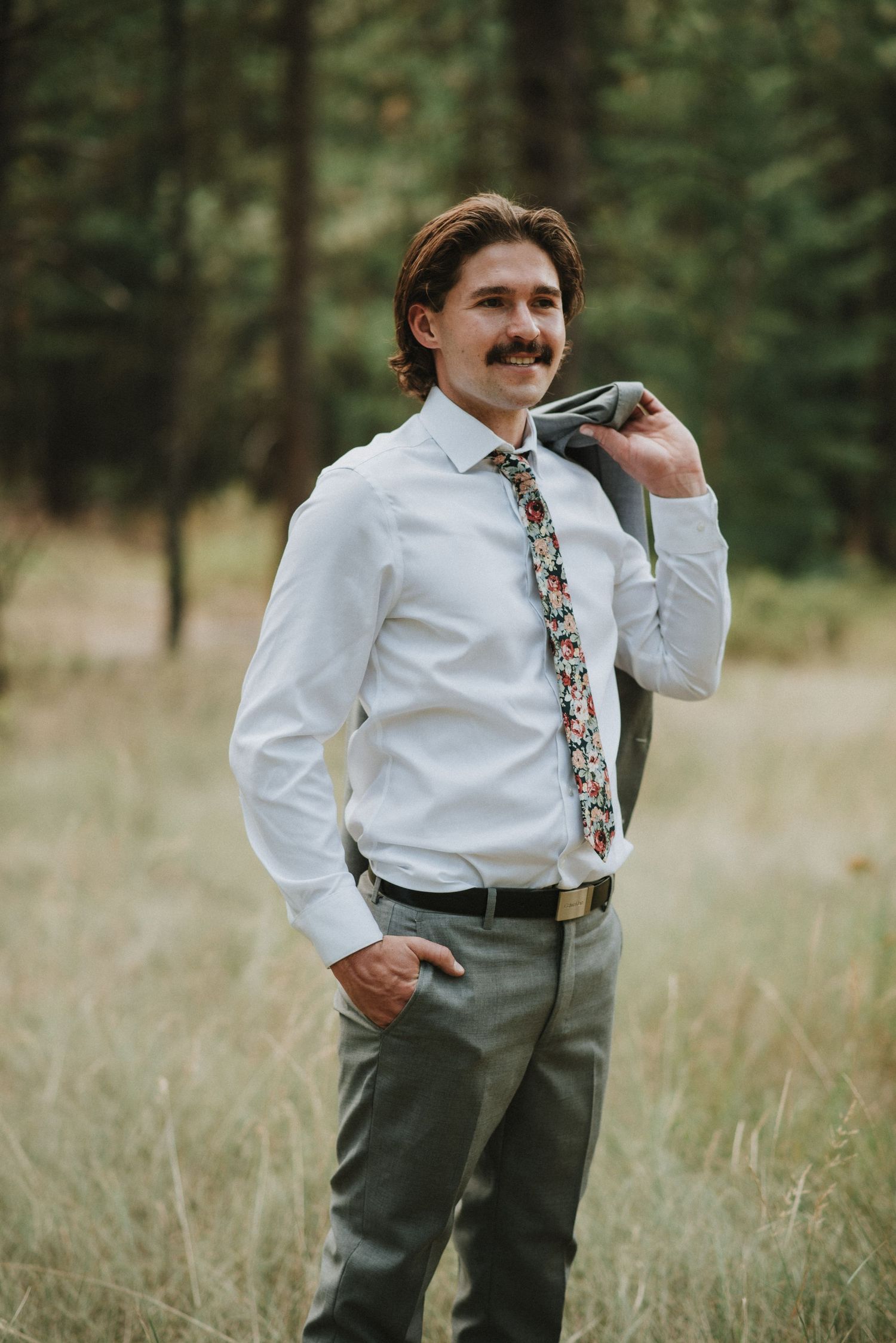 Picture of groom standing smiling. He is holding his suit jacket over his shoulder.
